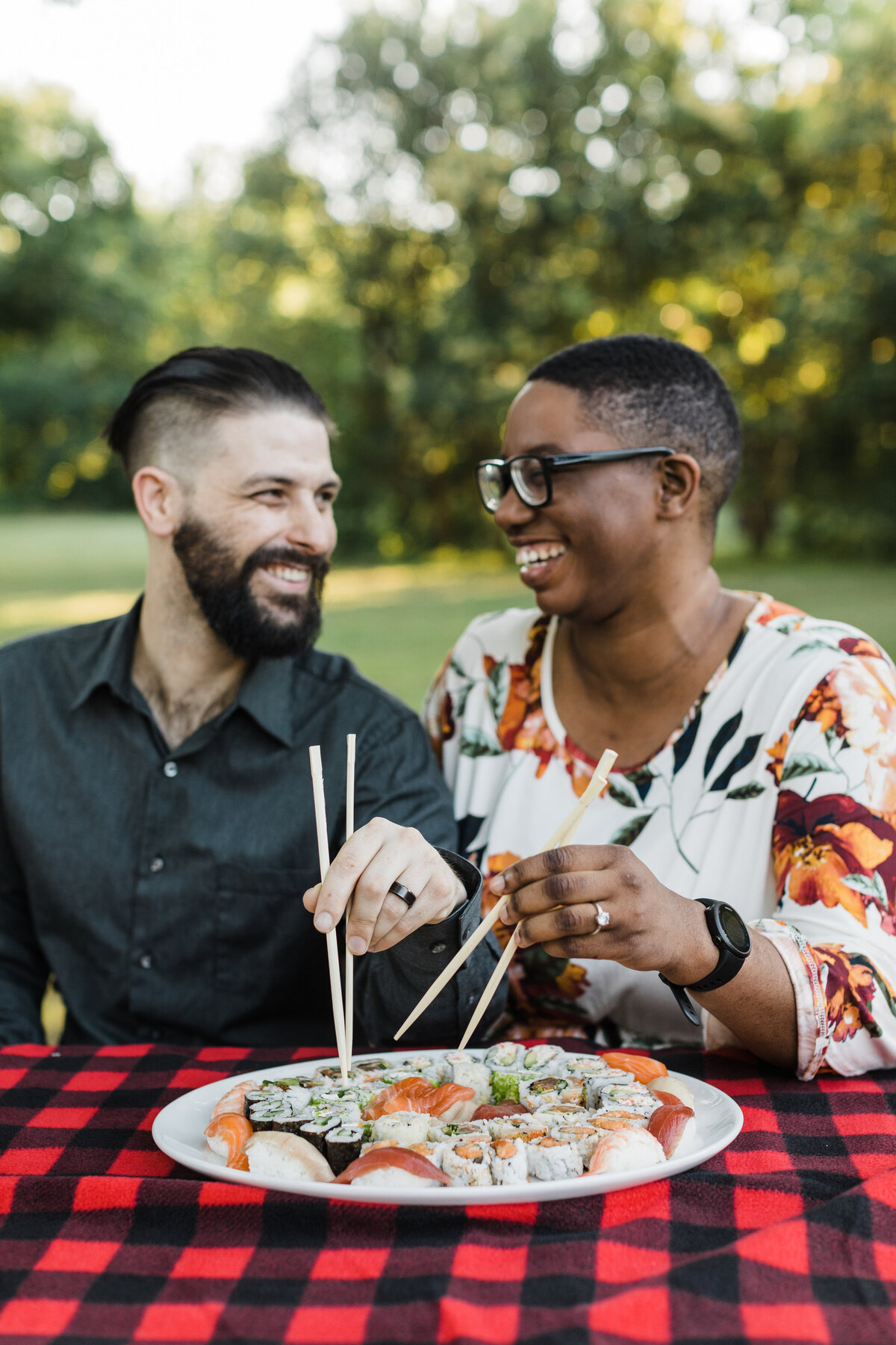 A couple smiling at each other while sharing a plate of sushi during their outdoor engagement session in Fort Worth, Texas. The woman on the right is wearing a white top covered in colorful flowers and glasses. The man on the left is wearing a dark button up shirt. The plate of sushi rests on a gingham tablecloth.