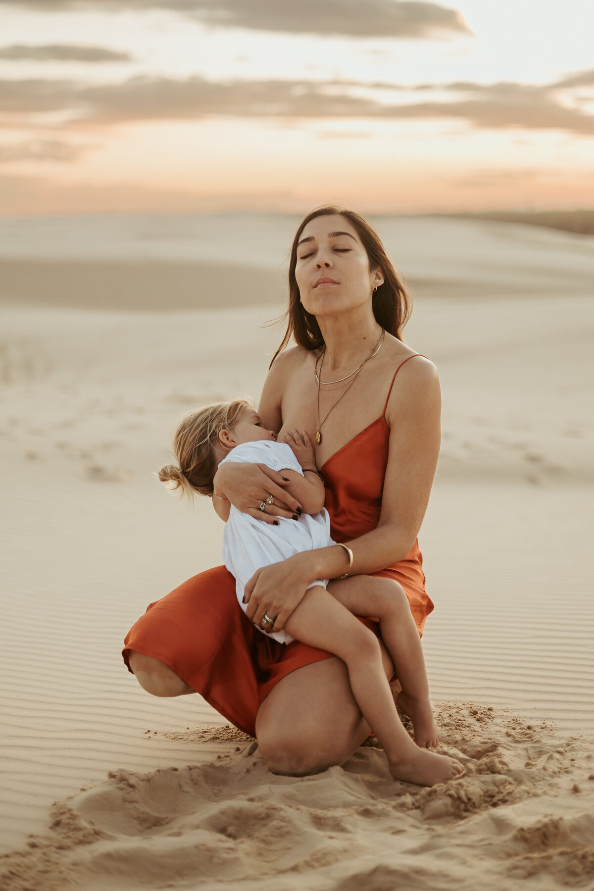 Mum in an orange dress with her eyes closed holding her daughter while she breastfeeds.