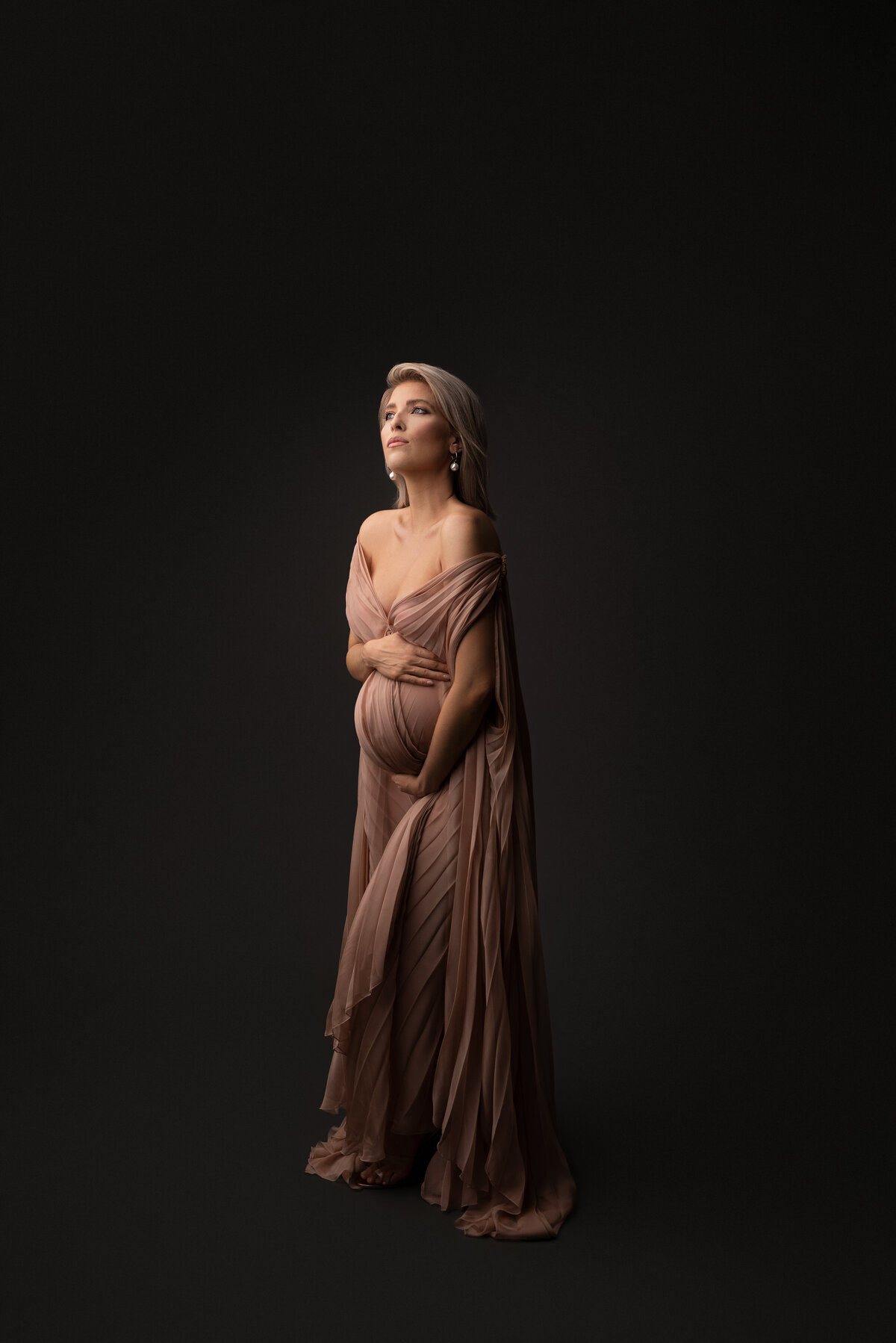 Woman poses for fine art maternity photos with New Jersey's best maternity photographer Katie Marshall. Full body image. Woman is standing in a long, dusty-rose floor-length gown with cape sleeves. One hand is under her bump, the other is over her bump. She is facing the light and looking upwards. The shadows to her right create depth to the photo.