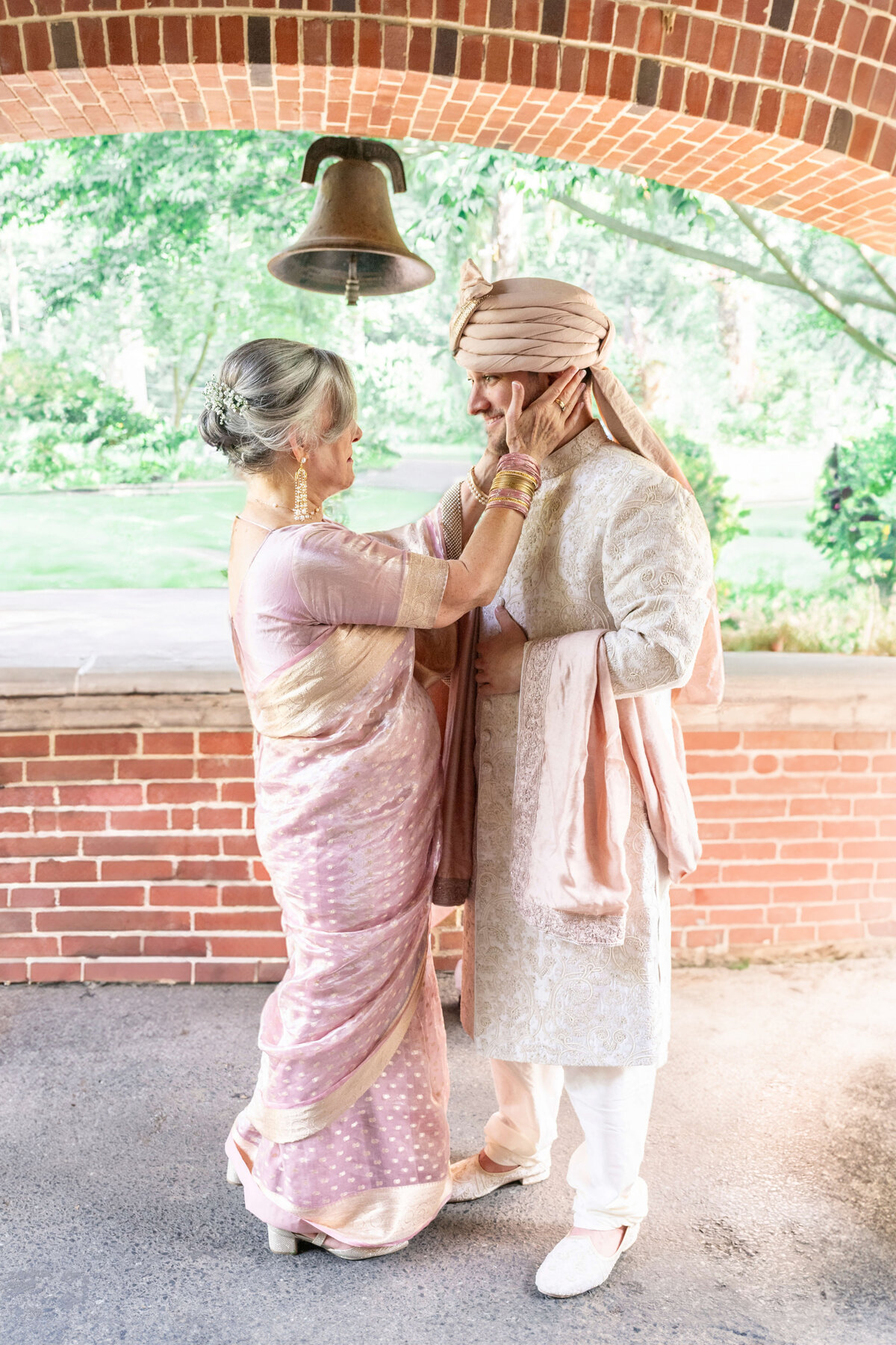 A mother and her son dressed in traditional Indian attire share an affectionate moment. The woman adjusts the mans turban under a brick archway, with lush greenery in the background.