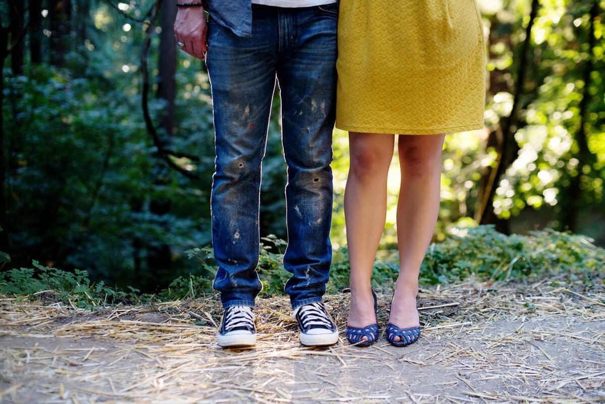 from the waist down, a woman is wearing a bright yellow skirt and blue shoes and a man wears paint covered jeans and converse