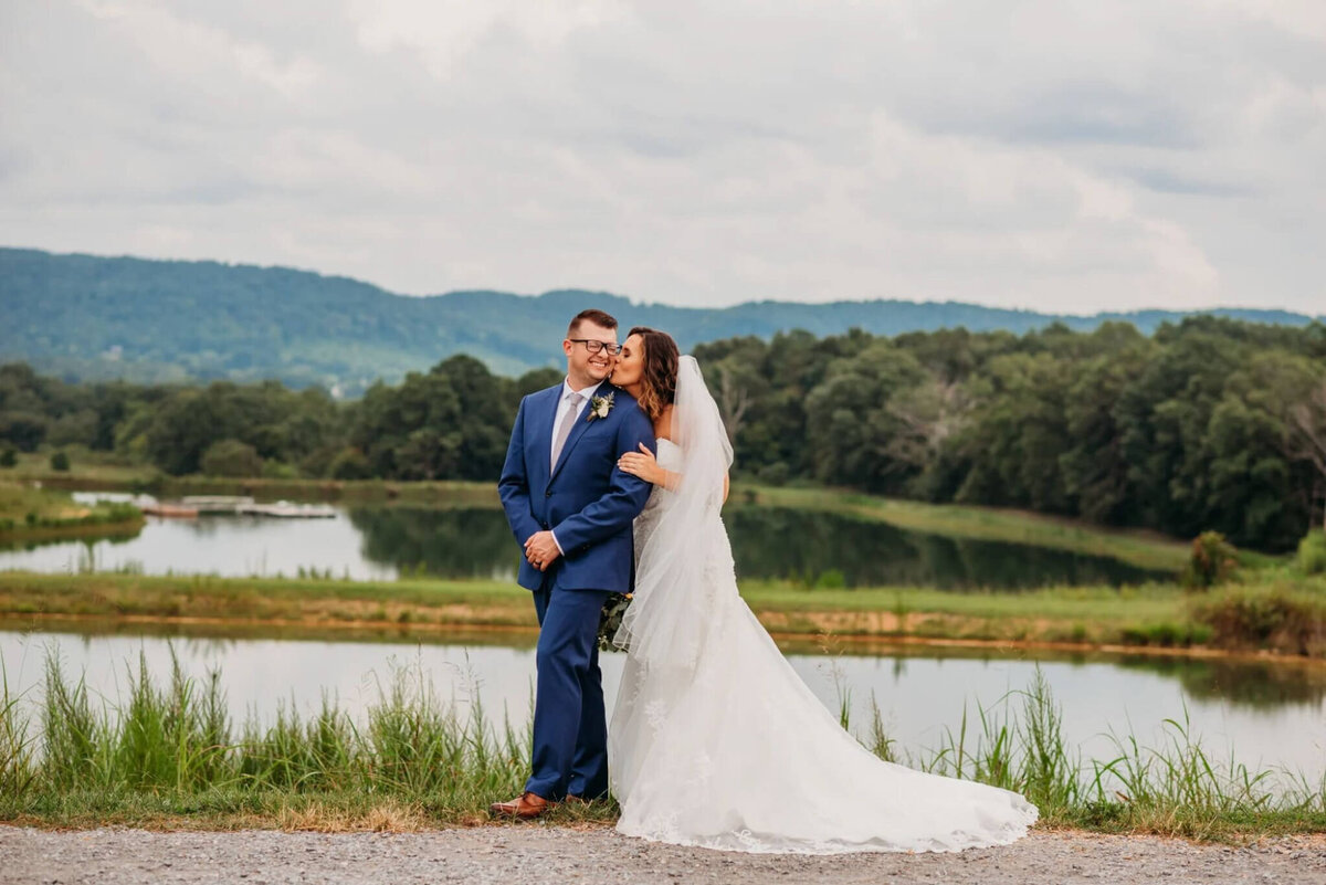 Photo of a bride cuddling his grooms arm and kissing his cheek while standing in front of a pond and mountains