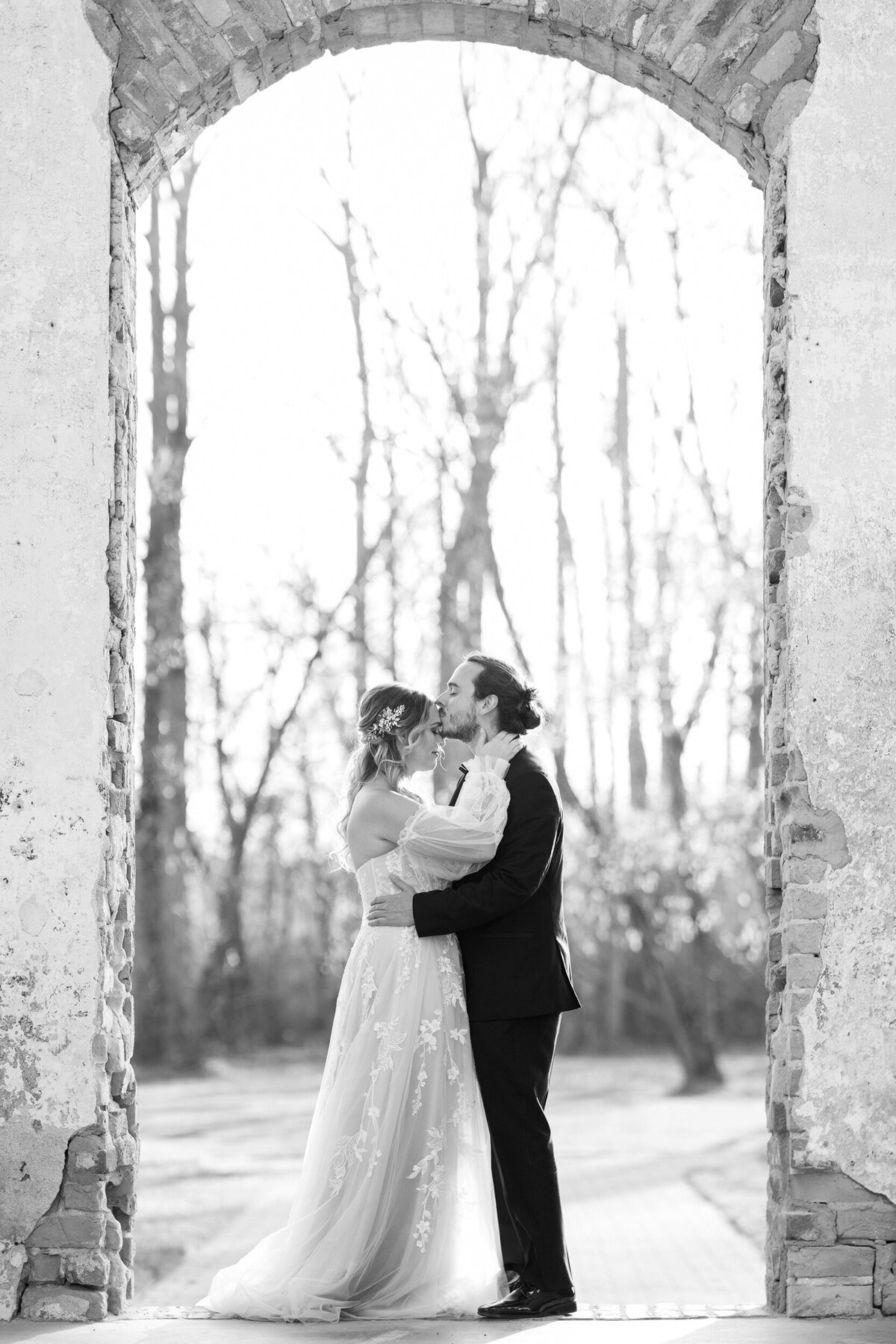 A black and white photo of a groom embracing and kissing his bride on her forehead in the doorway of the historic Providence Cotton Mill wedding venue near Charlotte, NC.
