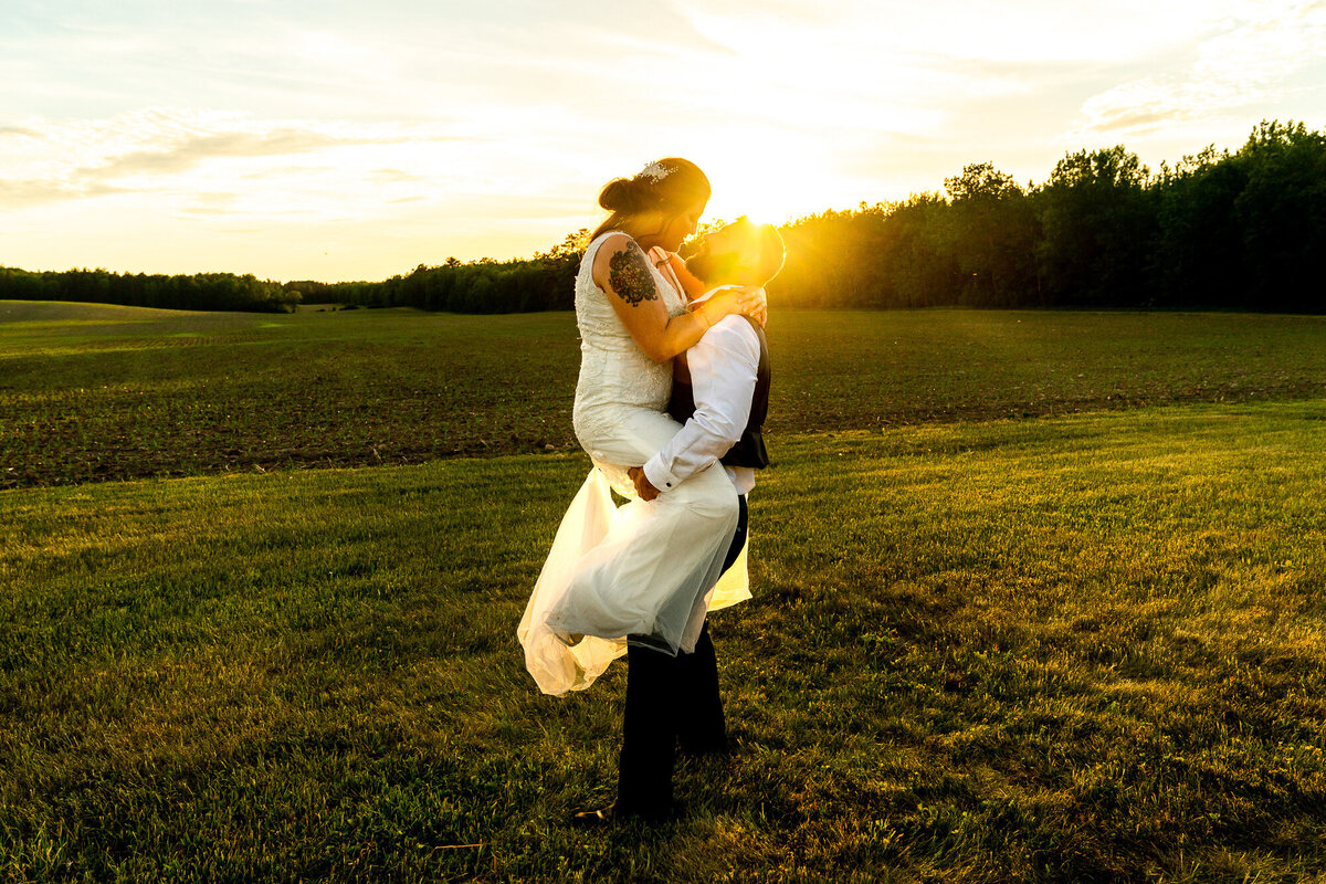 Groom lifts bride during sunset in na field.
