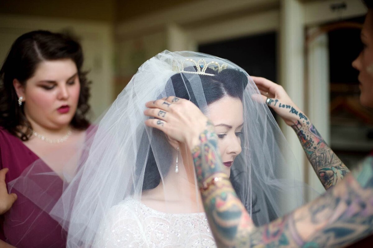 a bridesmaid with tattoo sleeves helps the bride put on her vintage veil
