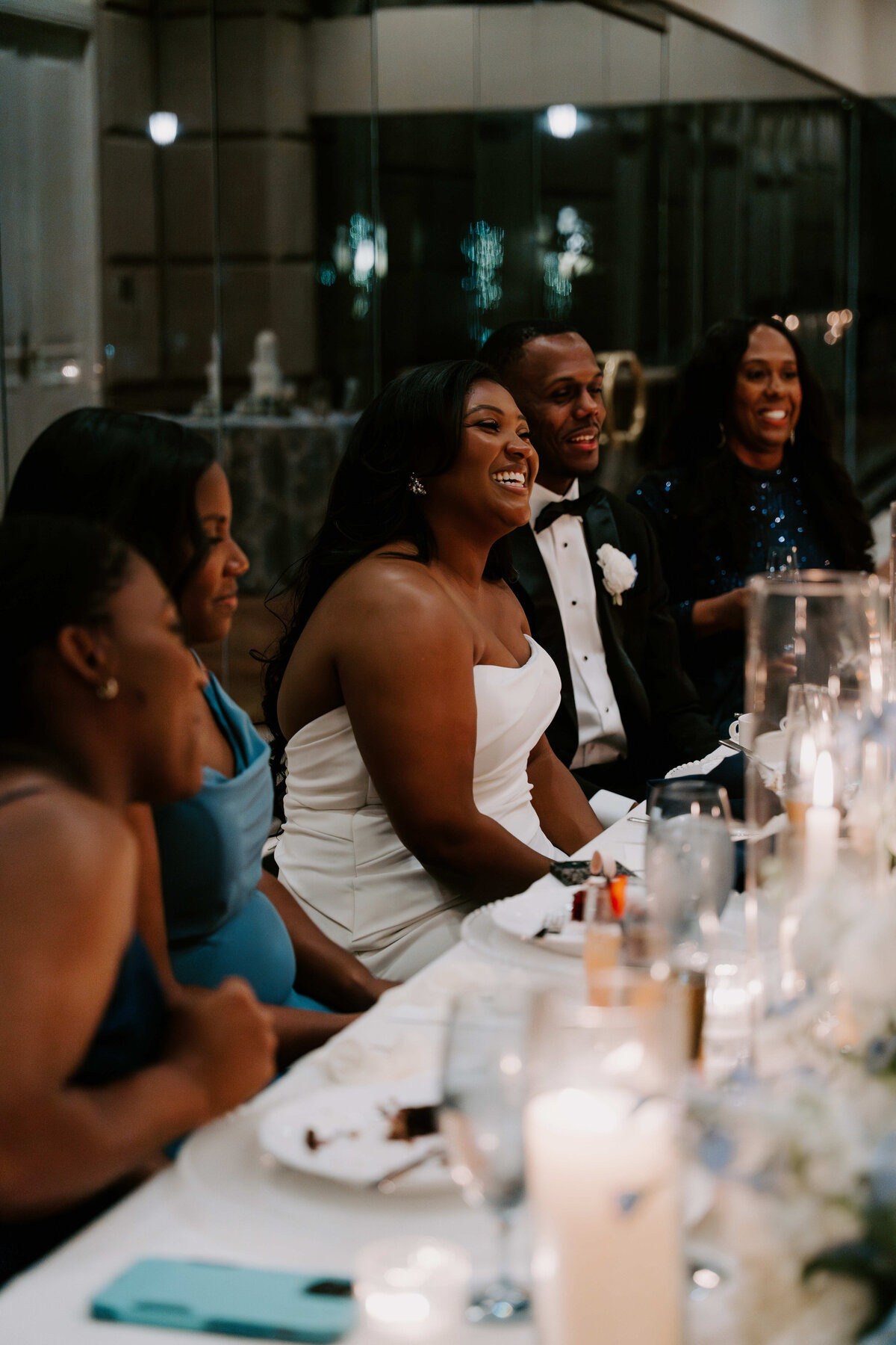 A joyful bride in a white dress and a groom in a tuxedo laugh with friends at an elegantly decorated table with candles and plates set for a wedding reception.