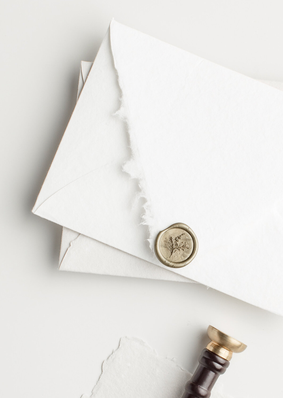 A stack of white handmade paper envelopes is shown, revealing the back flap  sealed with an antique gold wax seals. Beside the envelopes rests a wax stamp with a dark wood handle and brass stamp head.