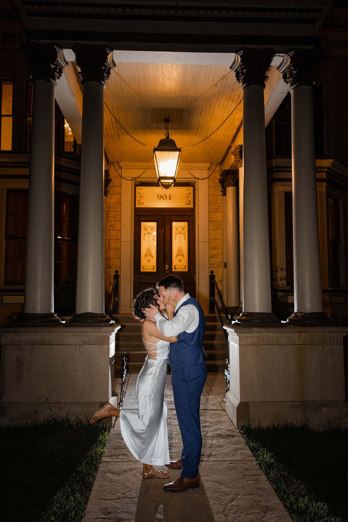 A couple embraces and kisses under a lantern at the entrance of a building with columns at night during their Iowa wedding.