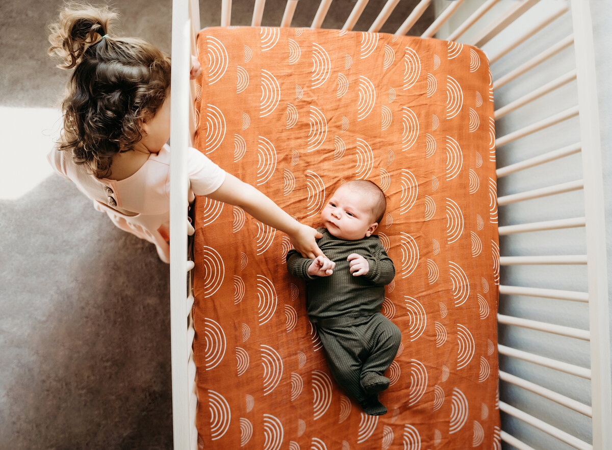 Newborn Photographer, a toddler sister reaches into the crib to admire baby brother
