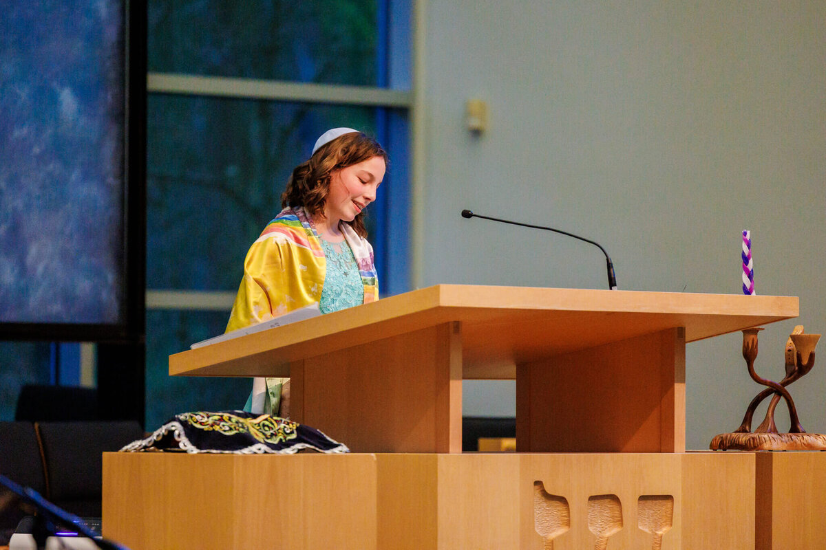 A girl smiles while standing at the bimah performing her haftarah in a rainbow tallit