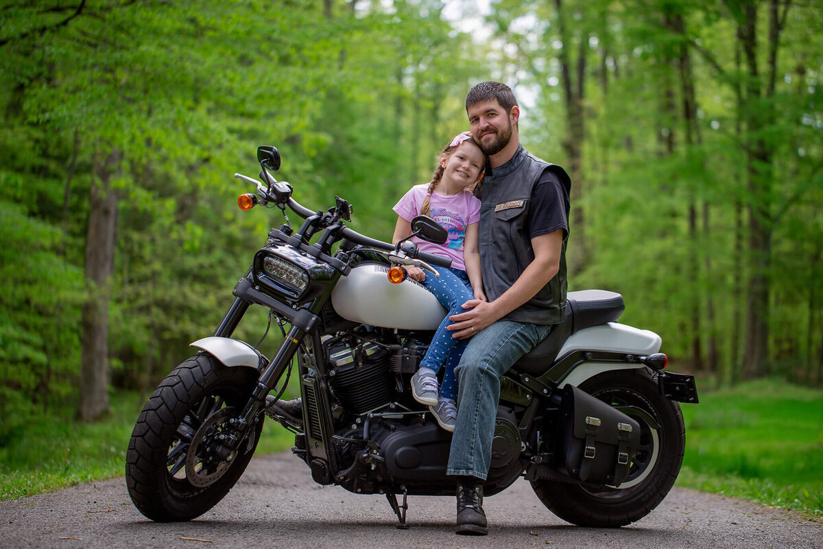 My husband and my daughter posing together on his Harley motorcycle with trees in the background.