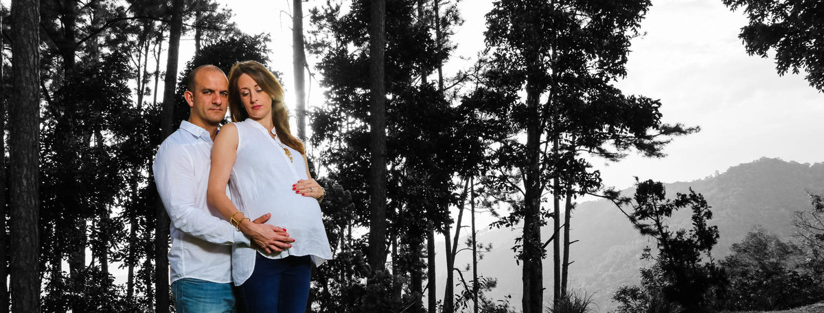 Pregnant mom and dad embrace in front of tall pine trees. By Ross Photography.