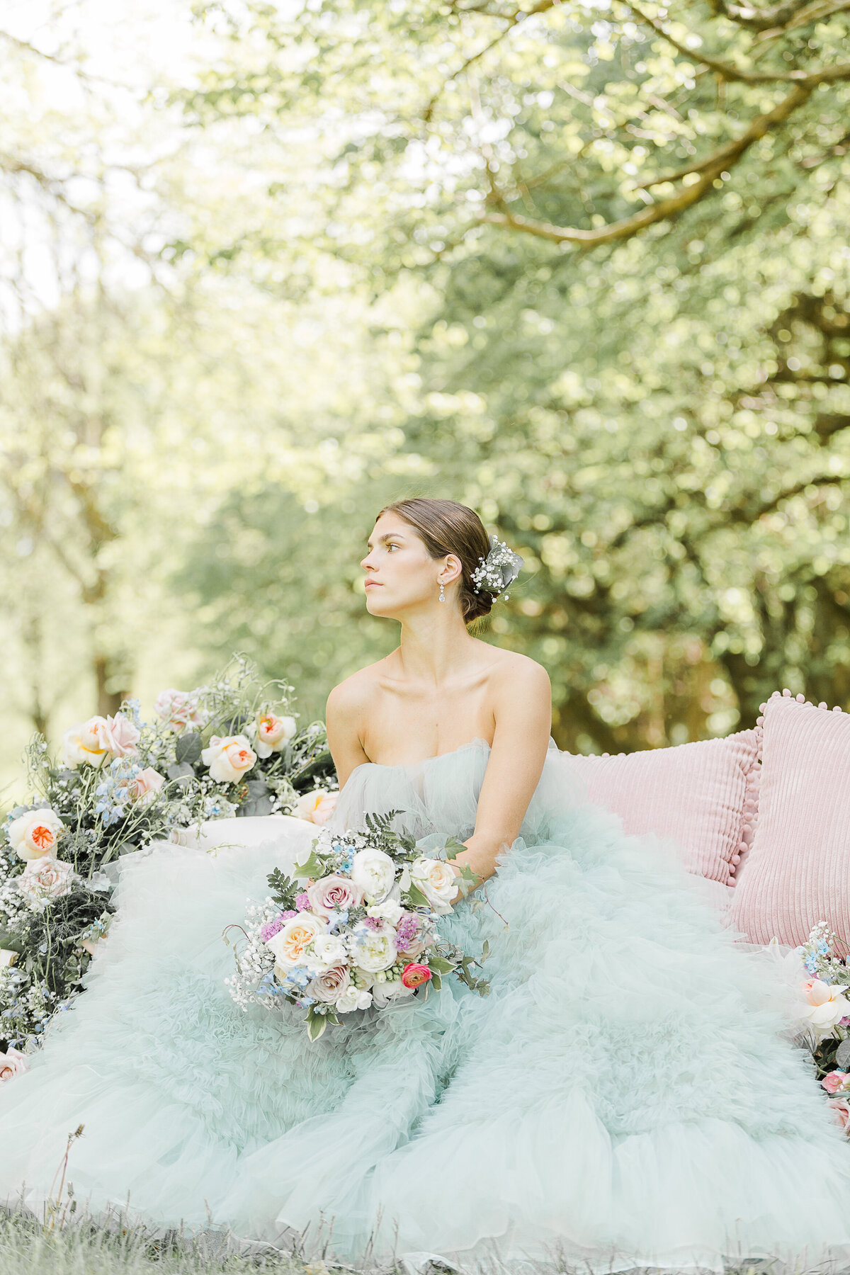 Bride poses for an editorial-style wedding portrait. Her gown is light blue and she is sitting on a pink couch outdoors and surrounded by bouquets of flowers. She is looking over her shoulder and into the distance. Captured by best New England Wedding Photographer Lia Rose Weddings