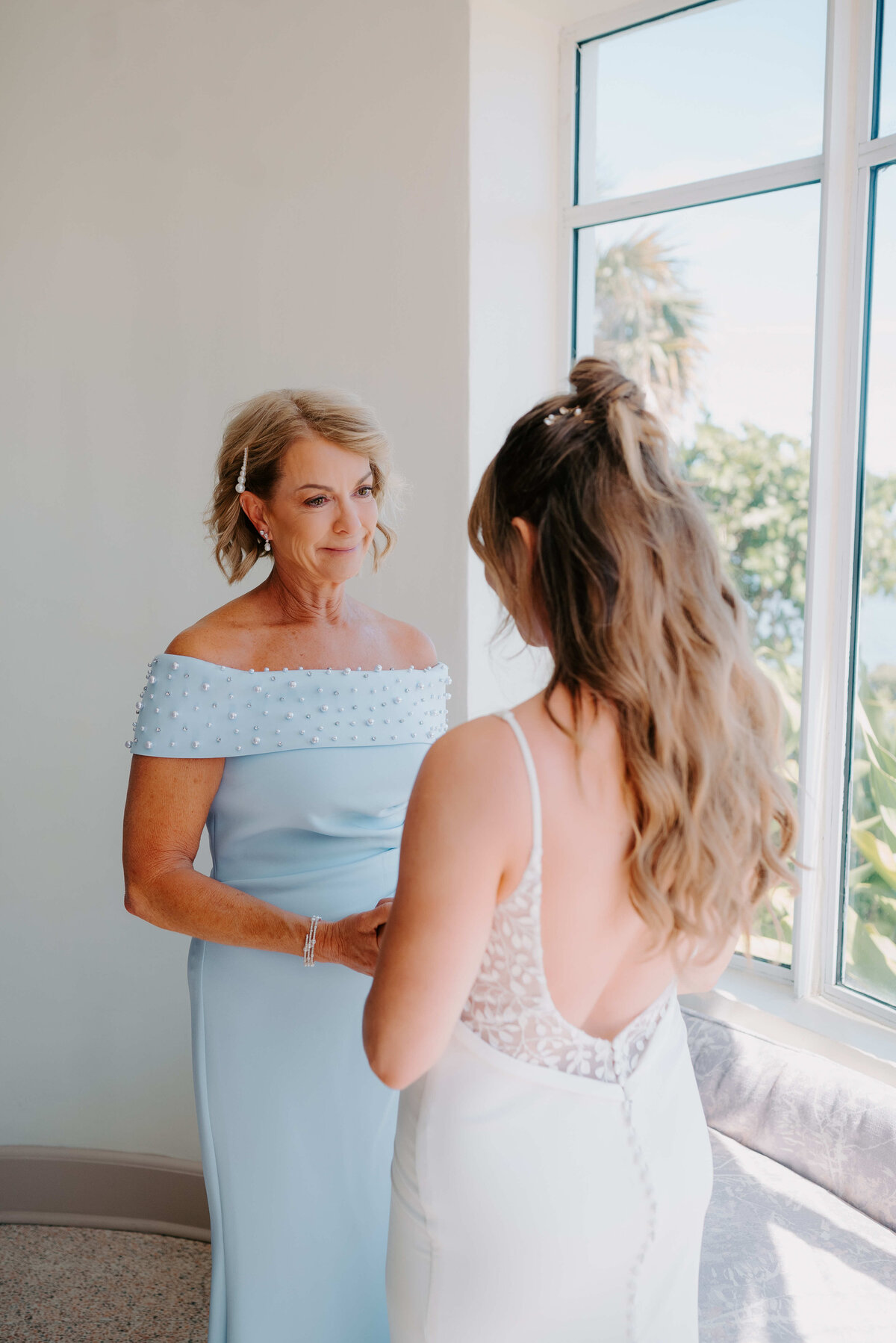 A woman in a light blue gown holds hands with a bride in a white dress, standing near a sunlit window. The scene conveys warmth and affection.
