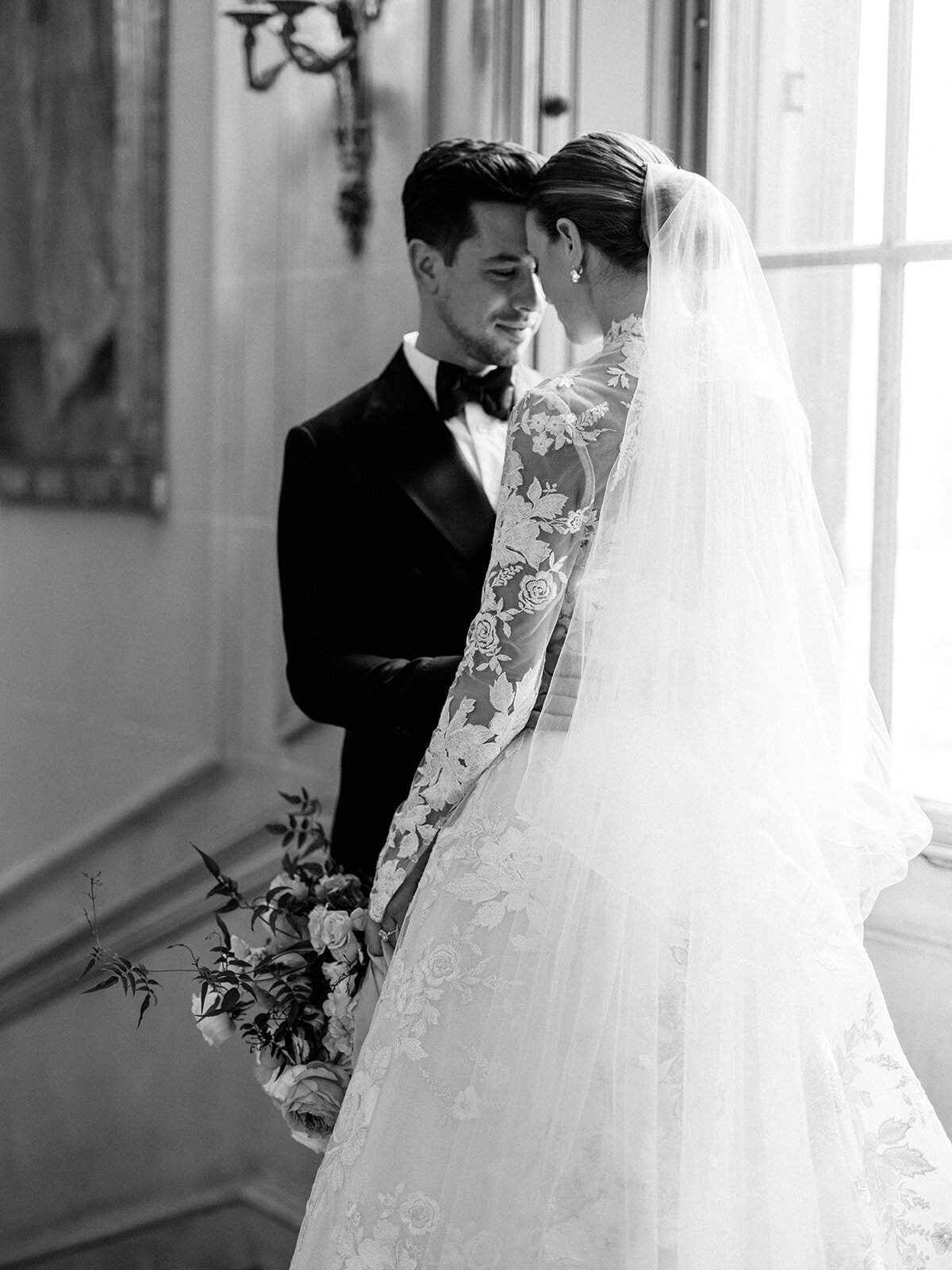 A black and white image of a couple by the window of Larz anderson wearing a tuxedo and gorgeous lace wedding gown.