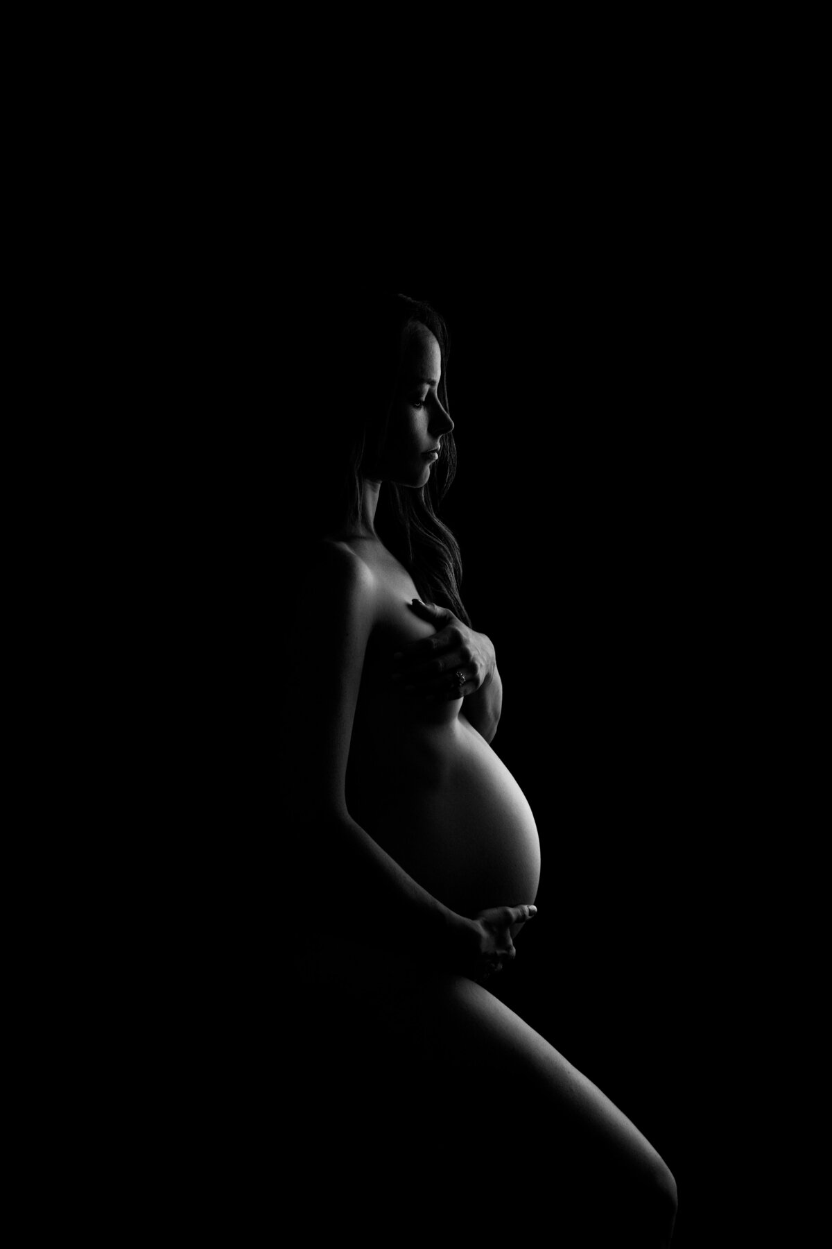 Philadelphia's premier maternity photographer, Katie Marshall, skillfully captures the drama and elegance of maternity in this striking black and white photograph. The expectant mother stands bare in side profile against a dark, dramatic backdrop.  With minimalist attire, she confidently showcases her pregnancy, her face in serene side profile. Soft light from the right gently caresses her form, creating subtle shadows that emphasize the contours of her body.  This captivating image artfully portrays the beauty and strength of maternity, evoking a sense of timeless grace and intrigue.