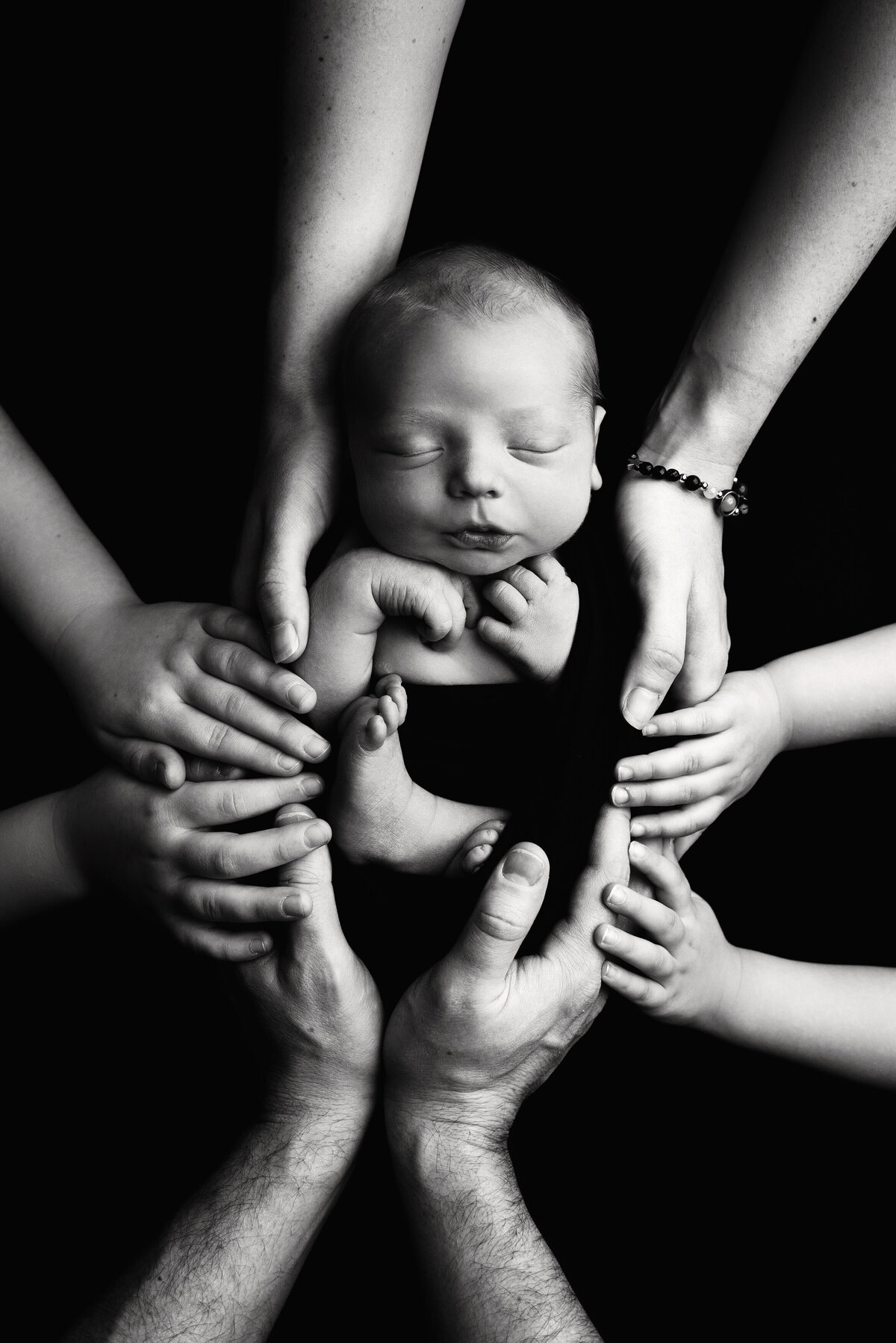 st-louis-newborn-photographer-black-and-white-photo-of-baby-cradled-by-four-sets-of-hands-wrapped-in-dark-cloth