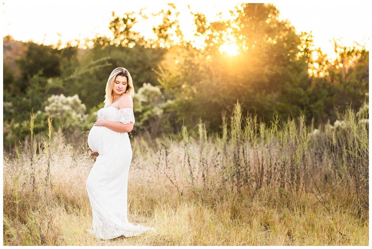 Woman in white lace maternity gown posing with hands on belly and looking down while standing in a field at Los Penasquitos Ranch House with the sun shining through the trees