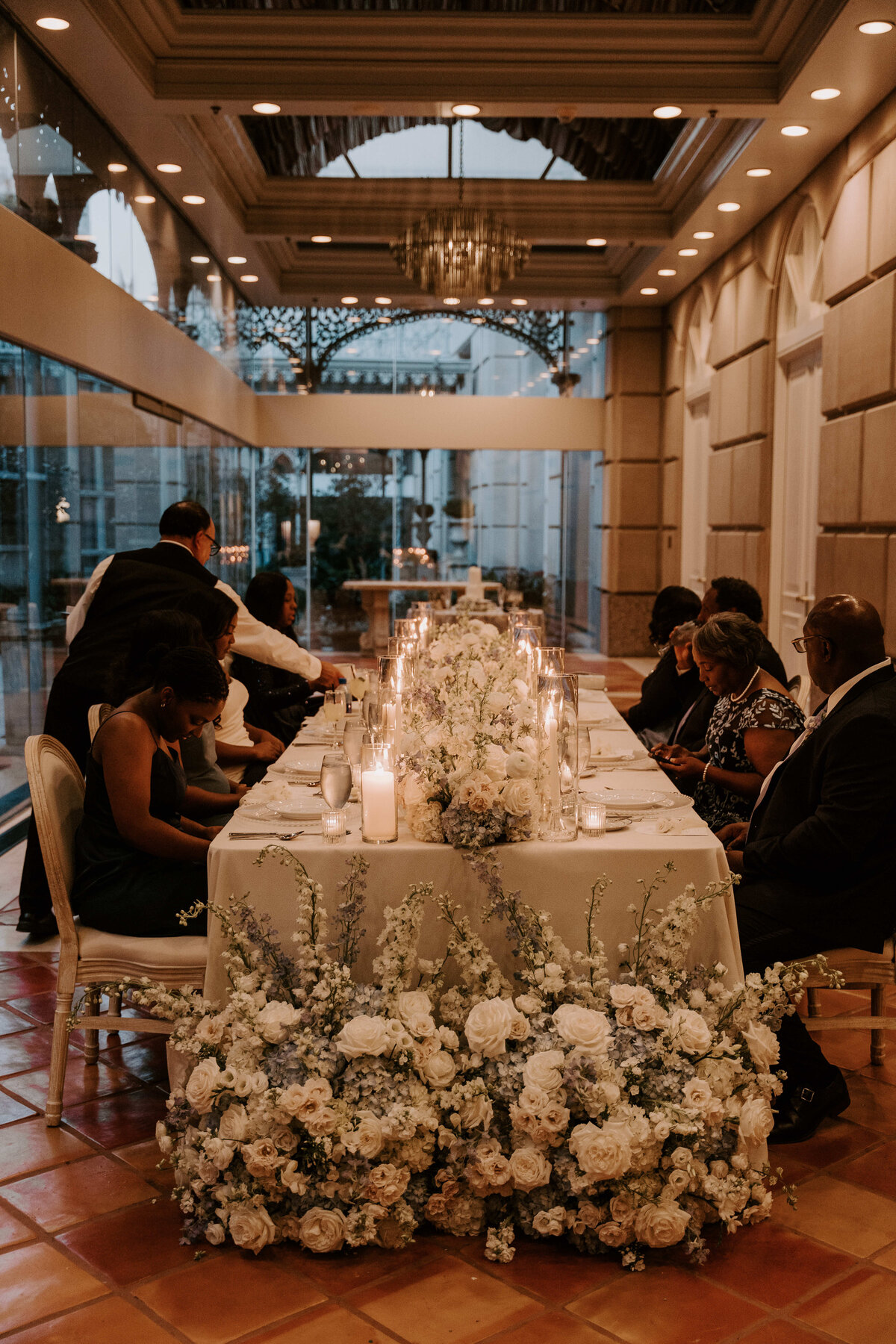 Elegant dining setting in a warm-toned room with people seated at a long table. White floral centerpiece, candles, and a cozy, intimate atmosphere.