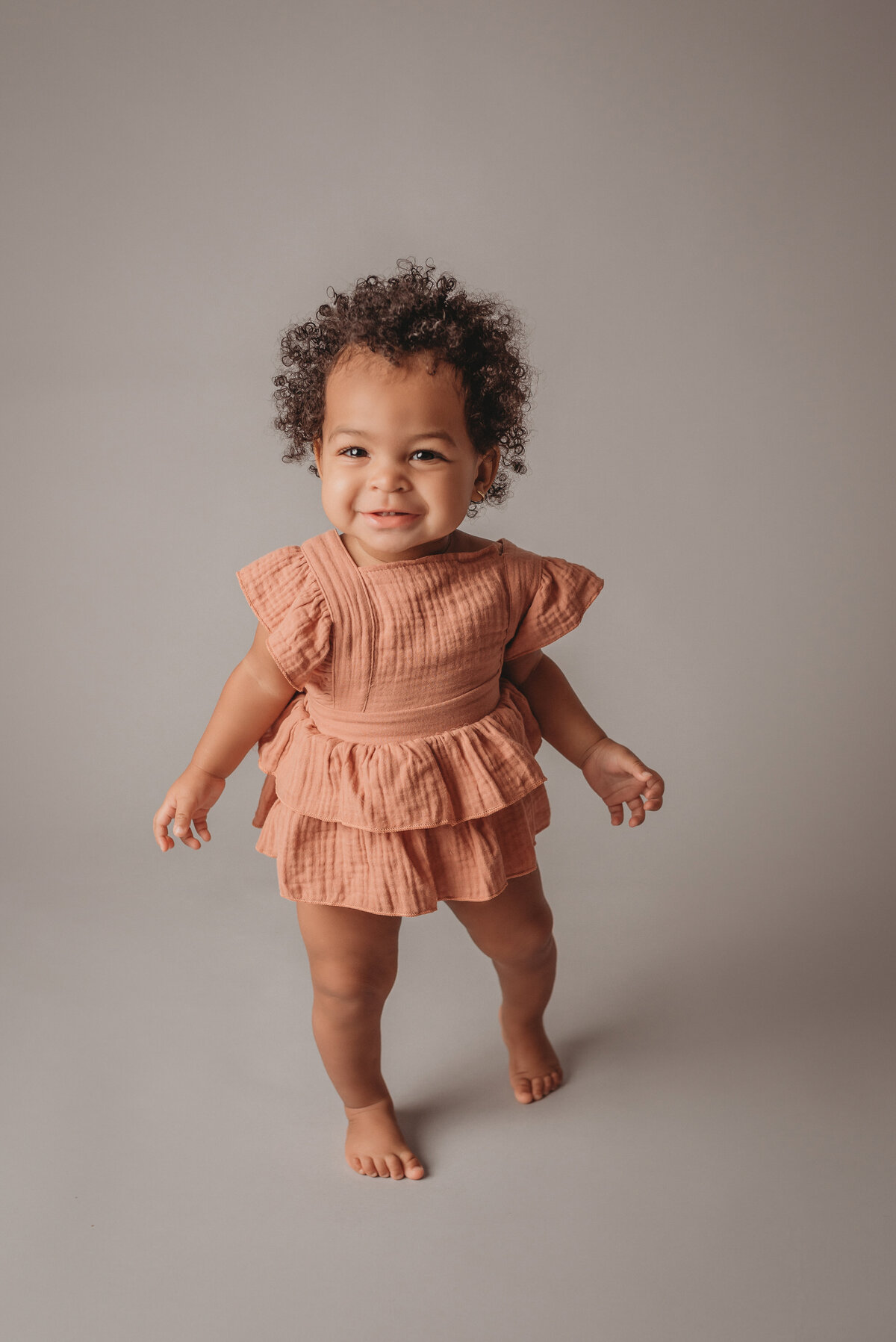One year old baby girl standing up wearing peach colored ruffle romper on light grey backdrop
