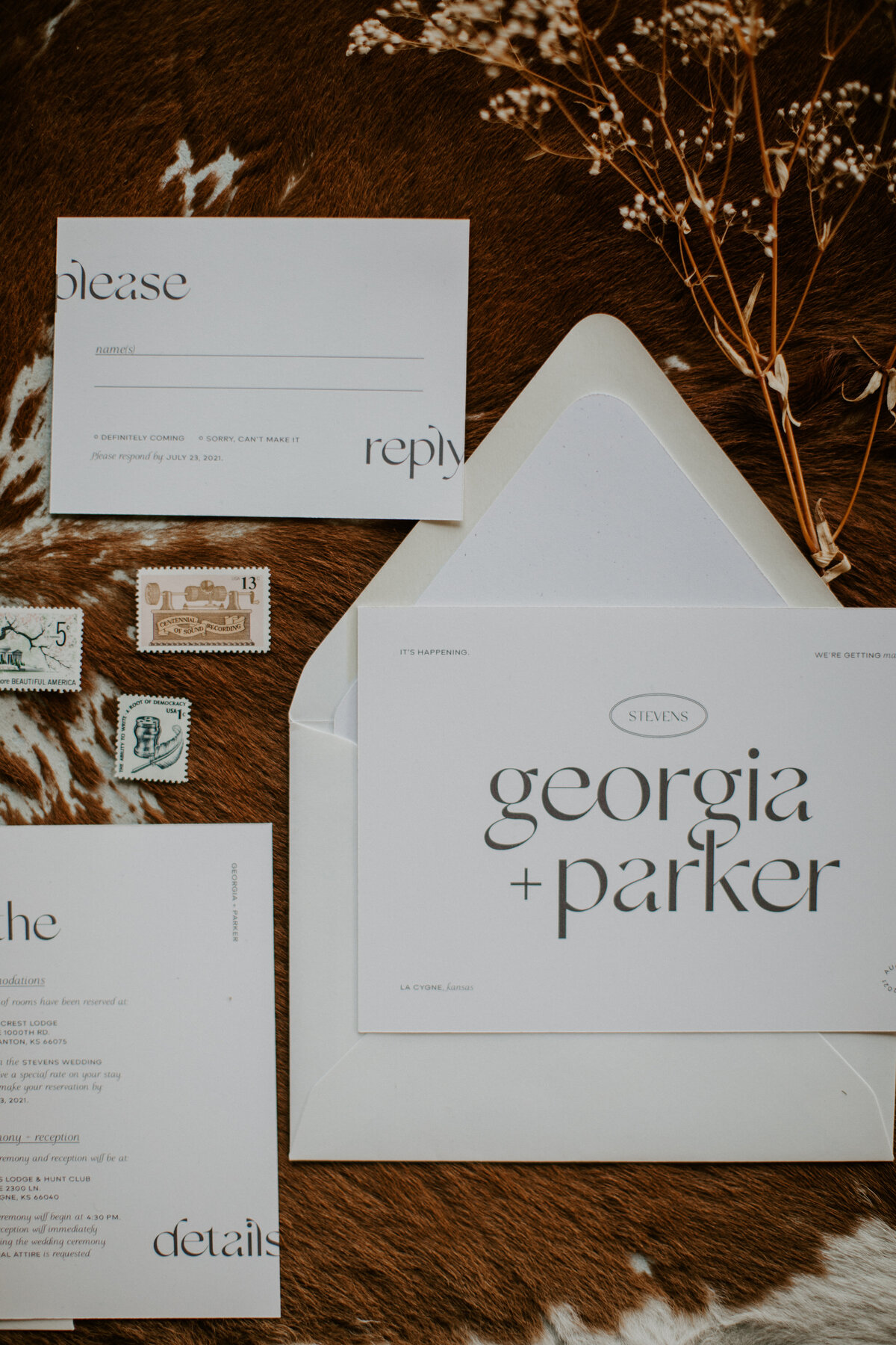 Various white wedding stationery with brown font and a dried flower stem set atop brown and white cow hide.