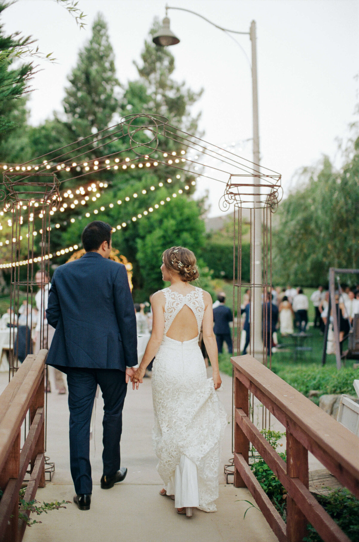 Husband and wife in their wedding attires walk past a narrow wooden bridge to join their guests for an outdoor reception party.