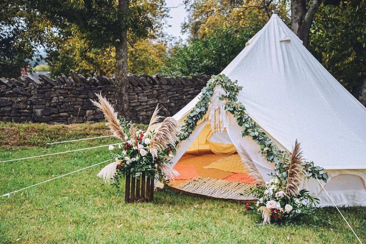 Teepee tent opening covered with greenery garland and flanked with two mixed flowers, greenery, and pampas