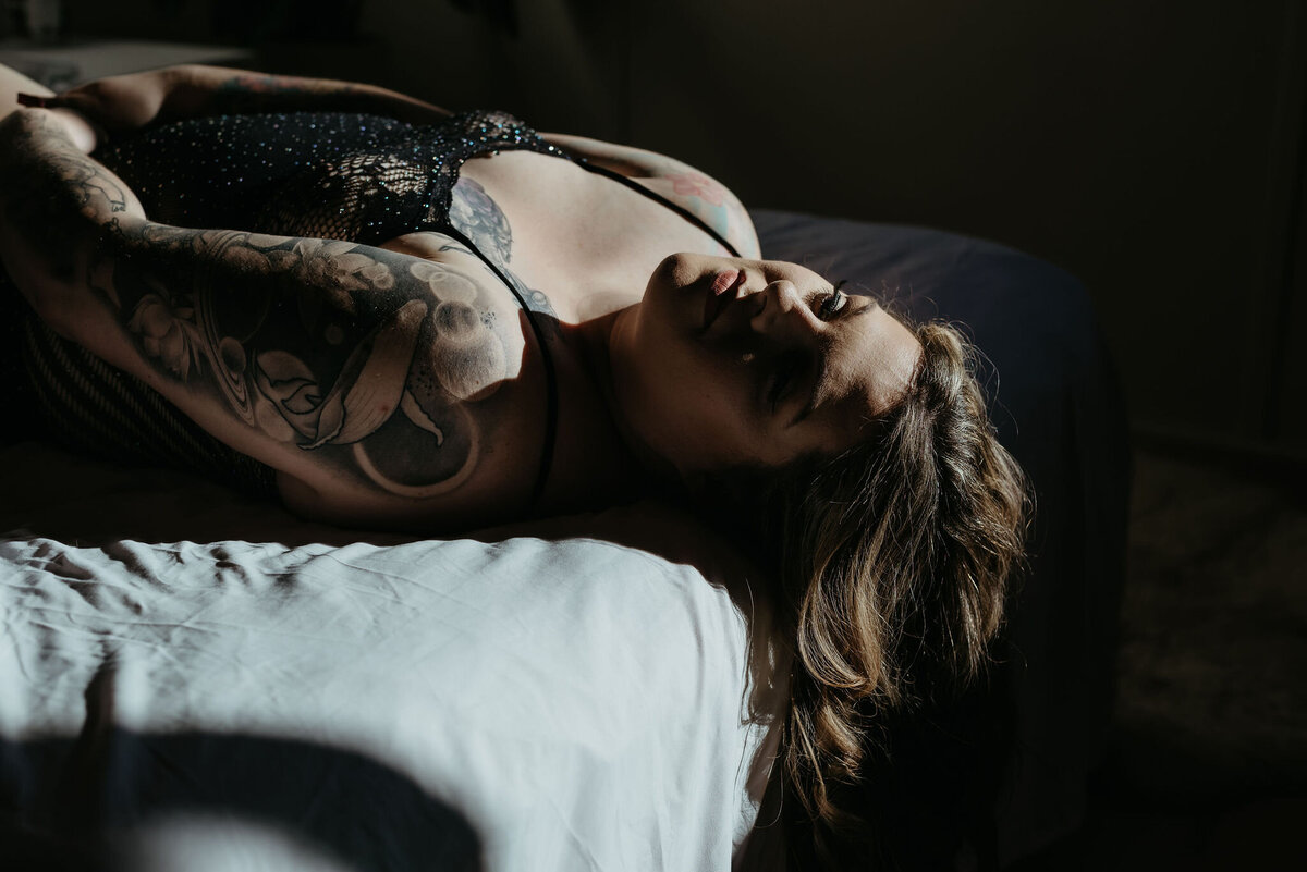 Woman in sparkly body suit lays on the bed in a patch of warm sunlight while posing for boudoir photos.