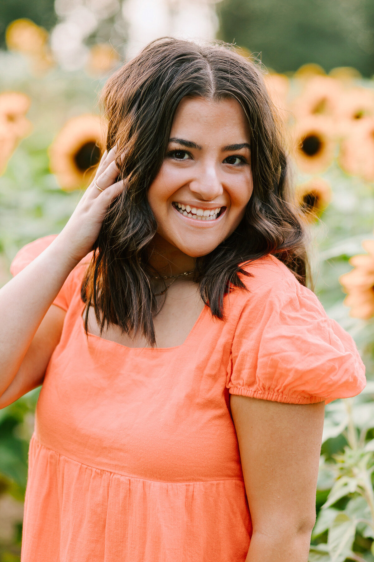 A teenage girl in a coral dress stands in a sunflower field laughing with her hand in her har