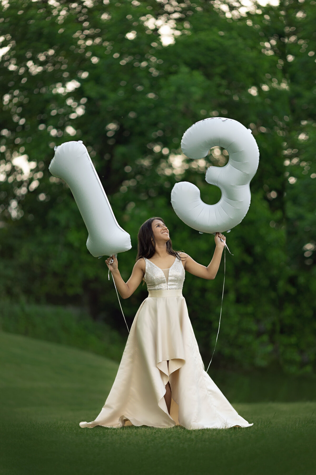 A teen girl in a cream dress holds a 1 and 3 balloon in a park