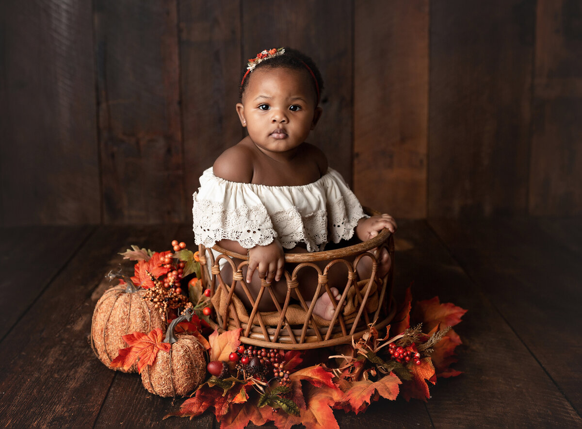 9-month baby girl photo shoot. Fall inspired. Baby is sitting in woven basket in a cream off the shoulder lace jumper and orange and cream headband. There are leaves, berries, and pumpkins at the base of the basket.