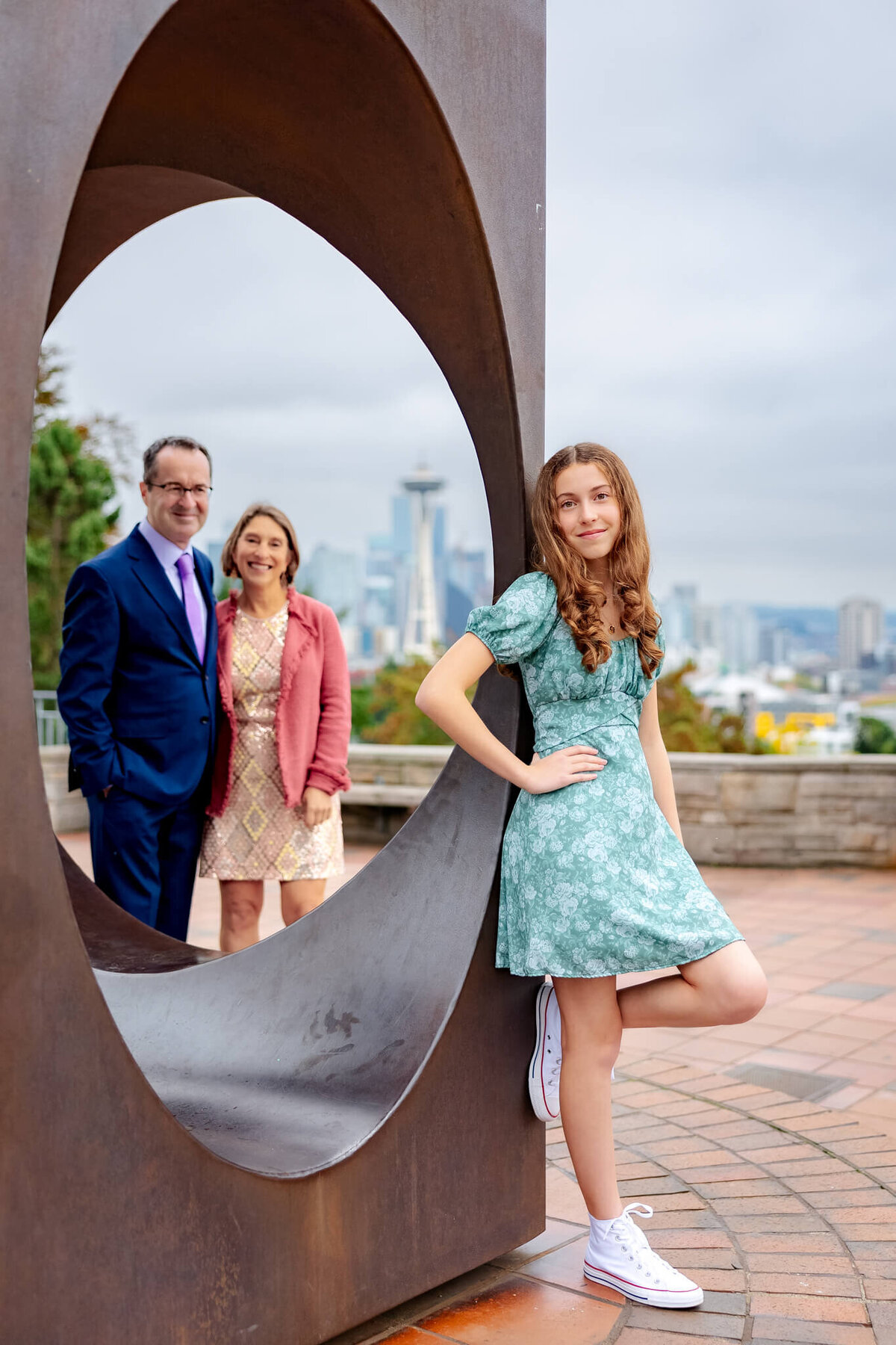 A teenage girl in a floral print dress leans against a sculpture in a park while mom and dad look on