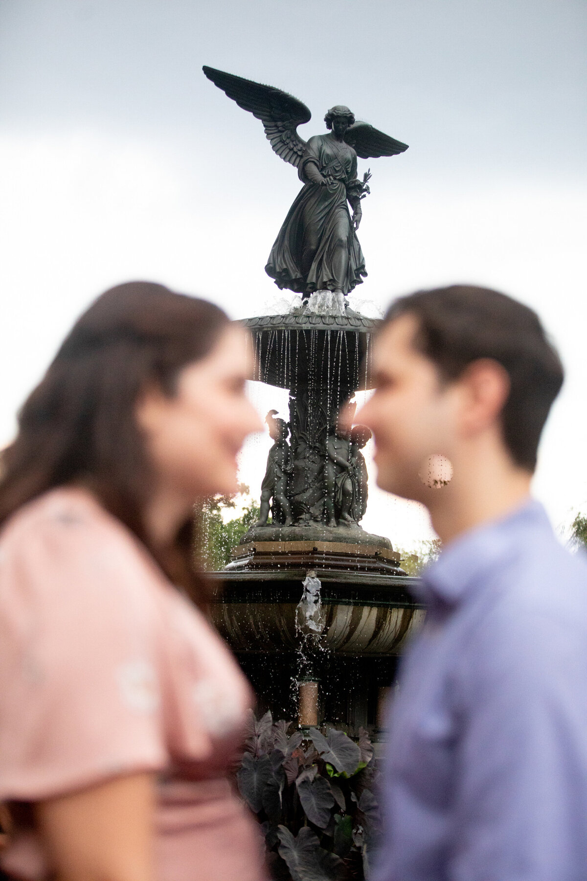A man in a blue dress shirt and jeans and a woman in a pink dress are out of focus with the angel fountain statue in focus between them in Central Park Bethesda Terrance New York wedding photographer