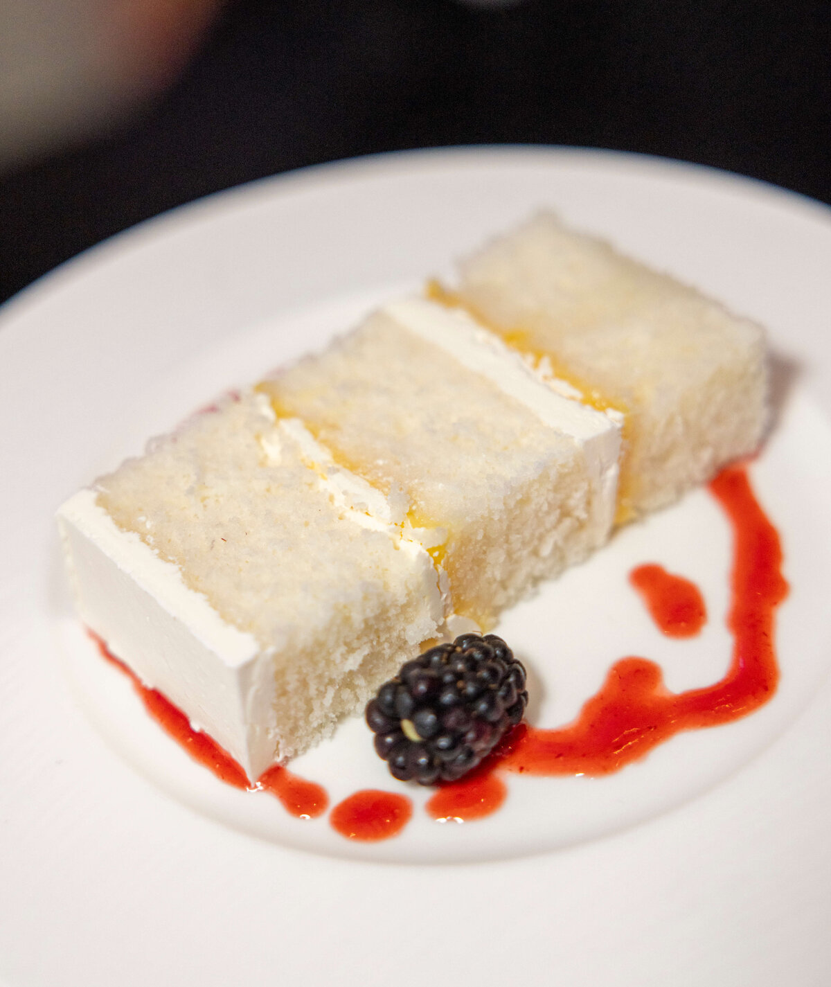 A slice of coconut cake with creamy frosting is arranged on a white plate, drizzled with a red sauce. A single blackberry accompanies the dessert, adding a touch of color.