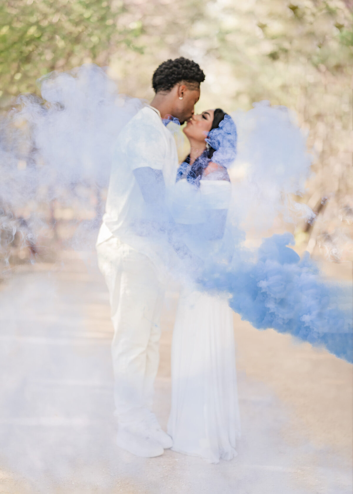 Expecting couple kisses behind gender reveal blue smoke bomb after learning they're having a boy during gender reveal photos at Brackenridge Park in San Antonio.