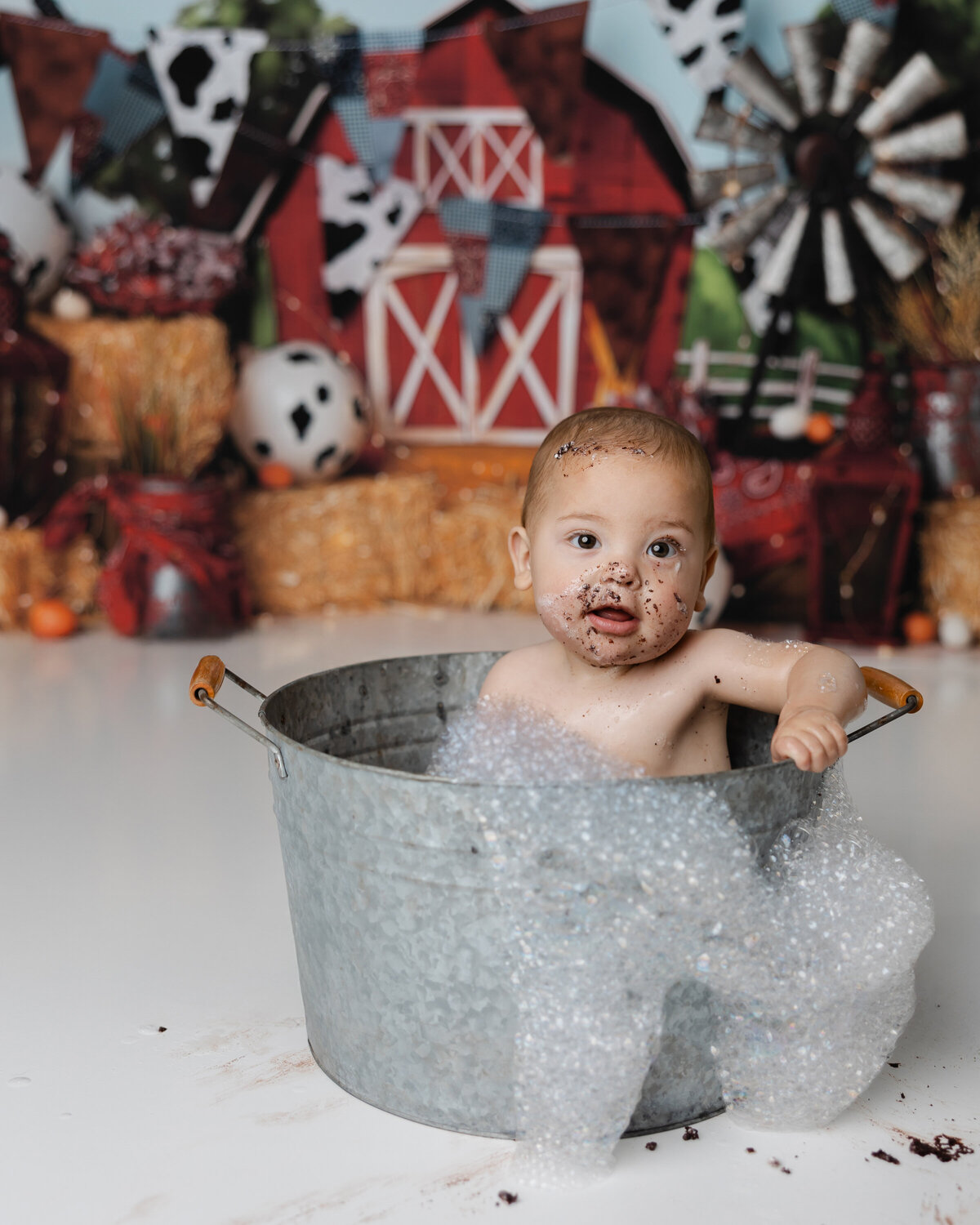 Farm-themed cake smash session ends with a cheerful bubble bath baby boy in a tin tub with a barn backdrop and frosting on his face.
Photograph by Yucaipa cake smash photographer Kaitlyn Dawn Photography.