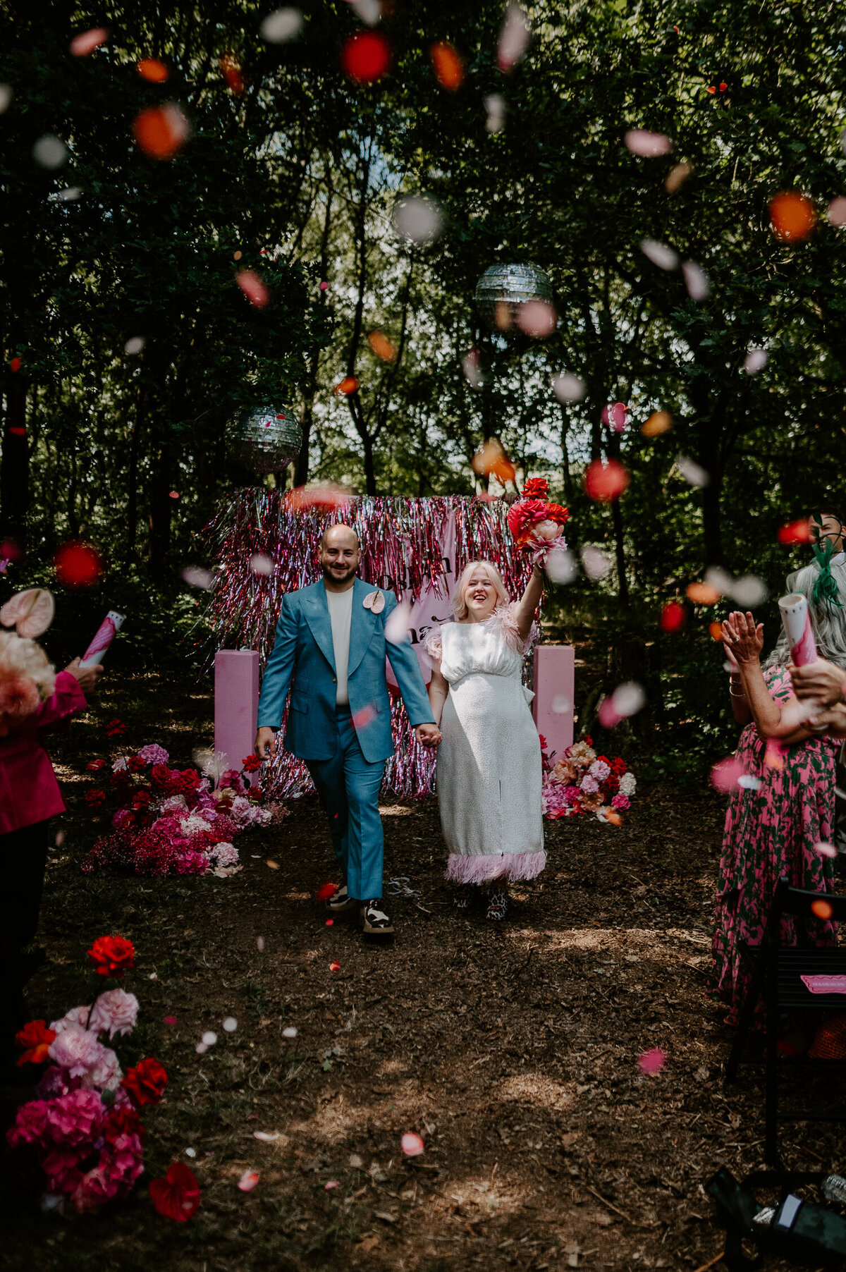 A Bride and Groom exiting their pink woodland wedding to their guests showering them with pink confetti. The bride has a short sleeve dress with pink fringing and holding a pink bouquet of flowers whilst the groom is wearing a blue suit with cow print Clarks Originals.