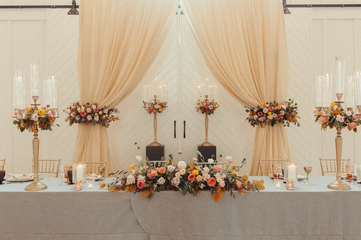 A long table is set with gray table cloths, flowers, and tall candlesticks against peach-colored curtains.