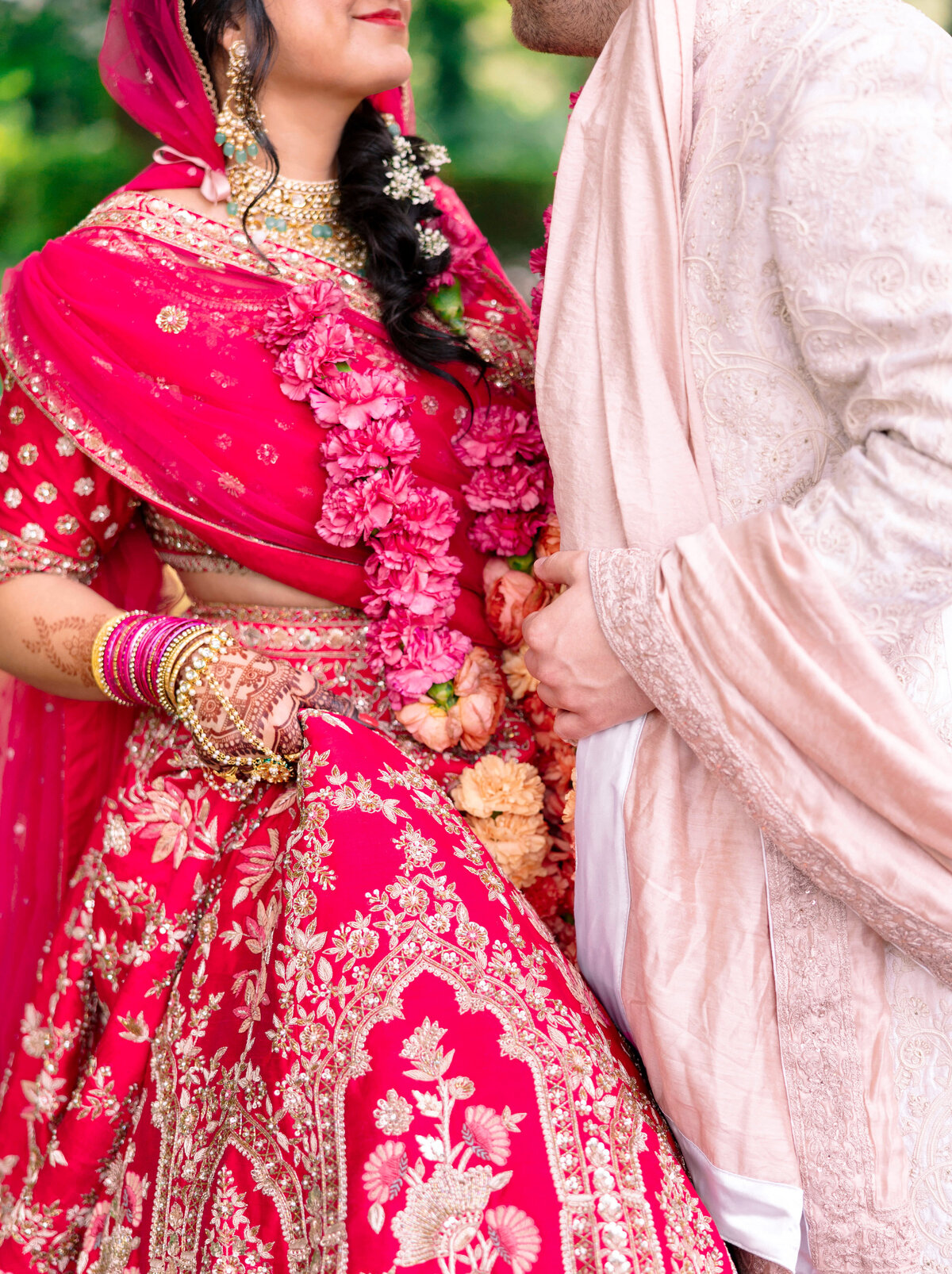 A couple in traditional attire faces each other. The woman wears a vibrant pink lehenga with intricate embroidery and gold jewelry. The man dons an ornate cream sherwani. Both wear floral garlands and are set against a blurred green background.