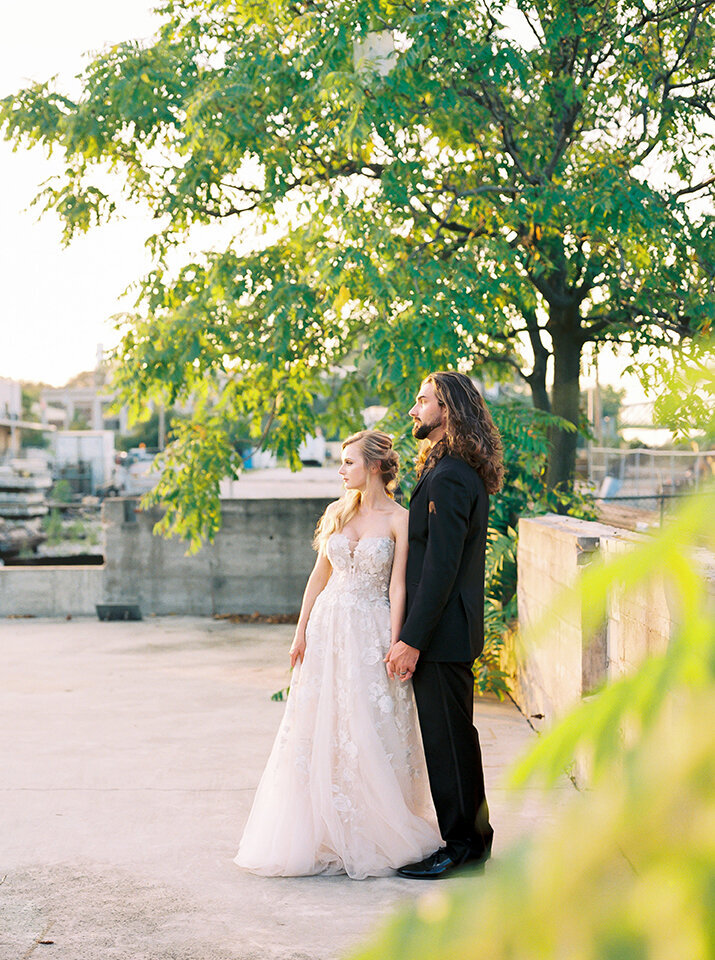 Side profile of bride and groom wearing a black tuxedo and white wedding gown holding hands on a rooftop.