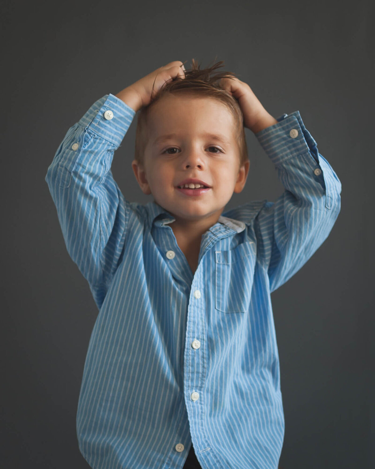 small boy putting hands on head in blue dress shirt with smirk on his face, with grey background