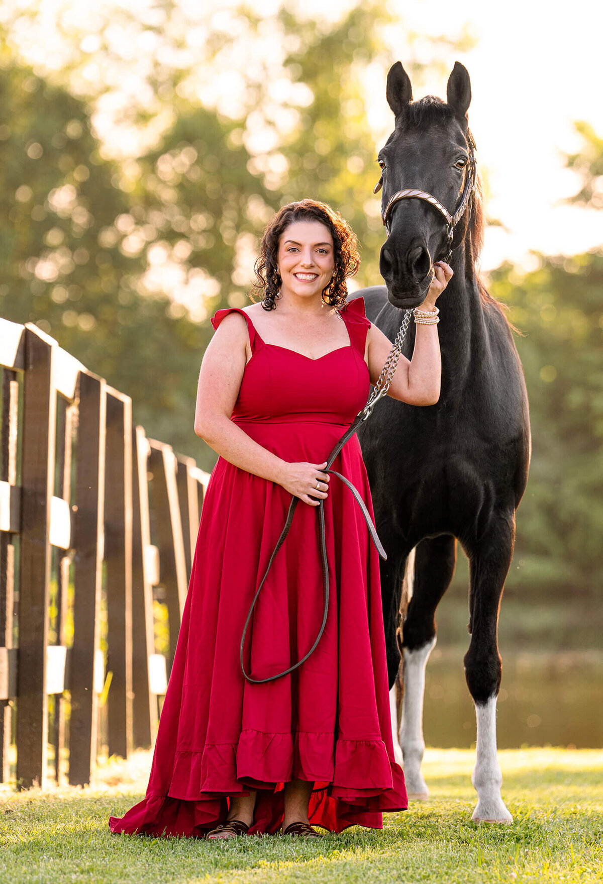 black haired female equestrian standing along a fence next to her black spotted saddle horse. behind them is a lake, trees, and beautiful golden hour sun.