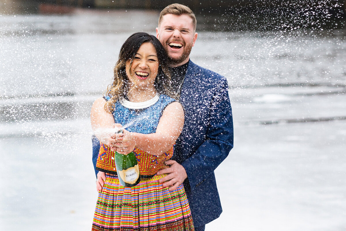 Asian woman and man spray champagne in front of ice covered river.