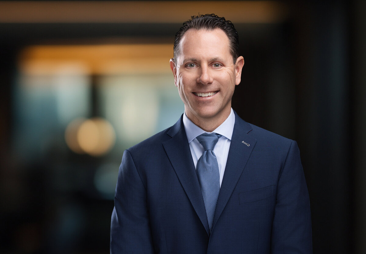 Professional headshot of a confident, approachable man in a navy blue suit and light blue tie, smiling with a genuine expression. The backdrop features a softly blurred, modern office environment with warm, ambient lighting, creating a polished, executive feel. Photographed by Janel Lee Photography, a Cincinnati-based portrait studio specializing in high-quality corporate headshots and personal branding photography for professionals in the Greater Cincinnati area.