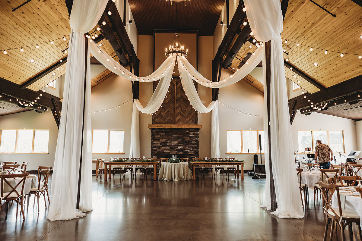 Mountain sage wedding reception decor at Camelot Ranch, featuring white drapery, ceiling lights, wood floors, wooden chairs, and elegantly decorated tables.