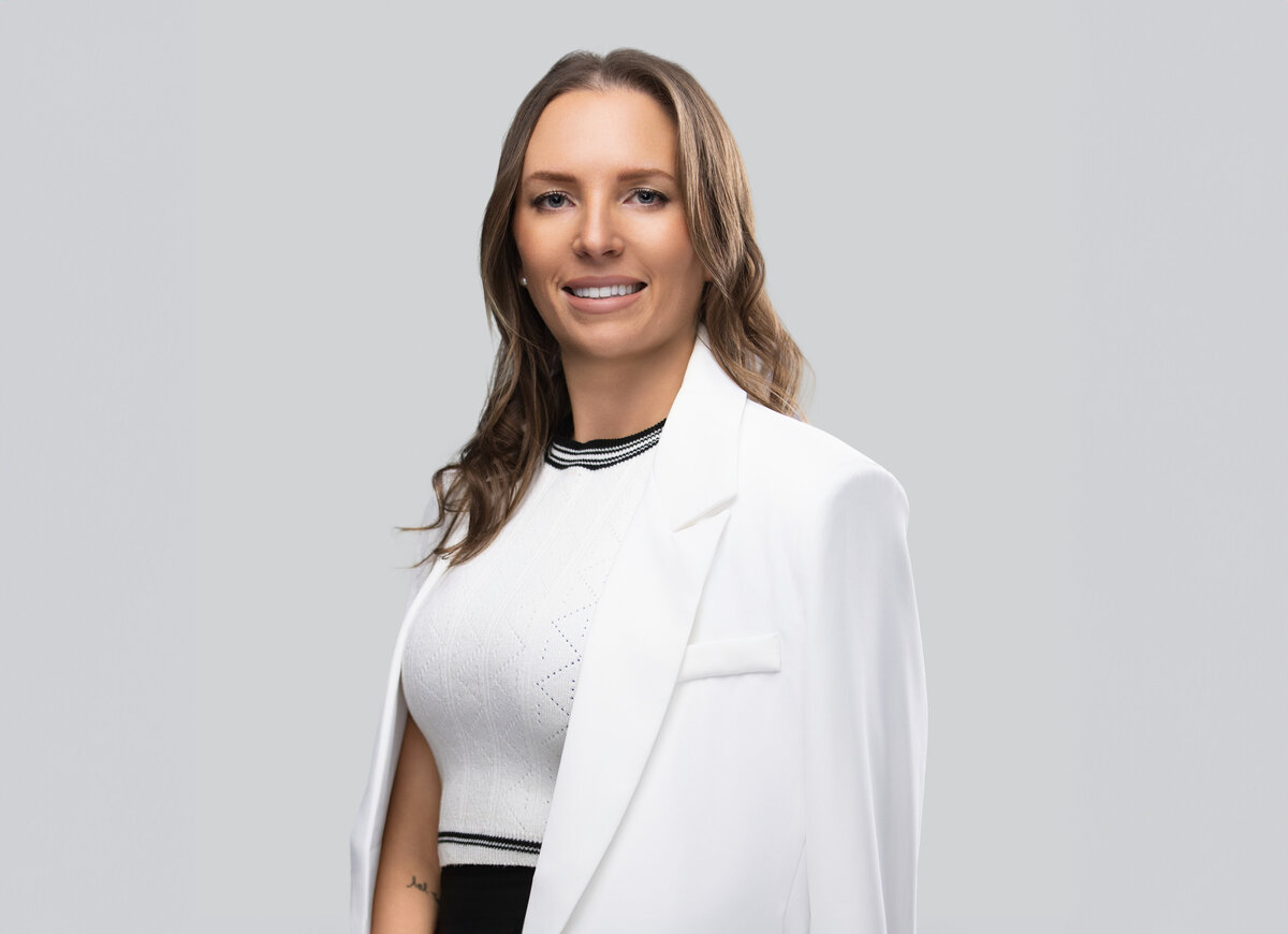 Professional headshot of a woman in Cincinnati, featuring a confident expression. She wears a tailored white blazer over a textured white top with black accents, paired with loose waves and a light gray background for a clean, modern look. Ideal for corporate and personal branding, this image highlights Cincinnati headshot photography at its finest.