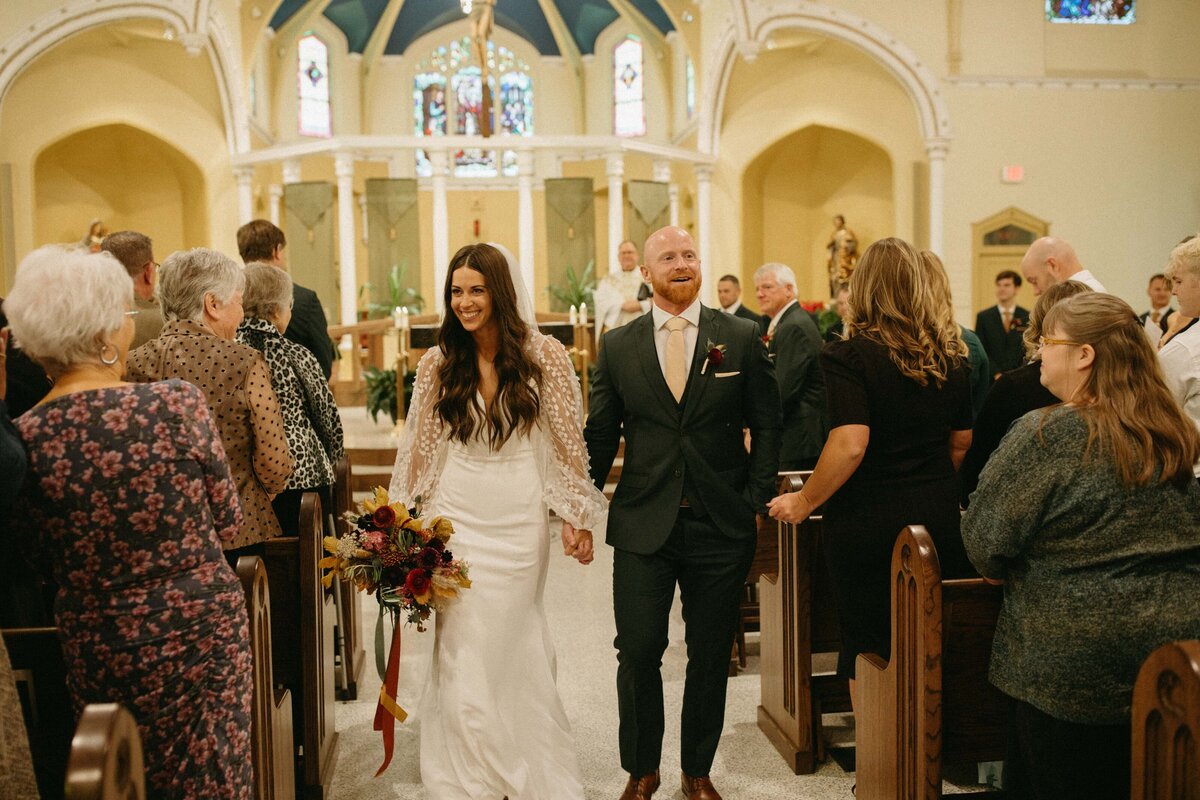 A bride and groom walk down the aisle, smiling, as guests look on in a church with stained glass windows and ornate arches, all meticulously orchestrated by a top wedding planner from Des Moines.