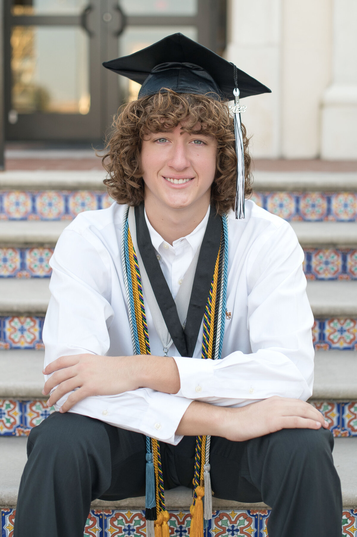 High school senior boy sitting on steps with cap and honors cords smiling at the camera.