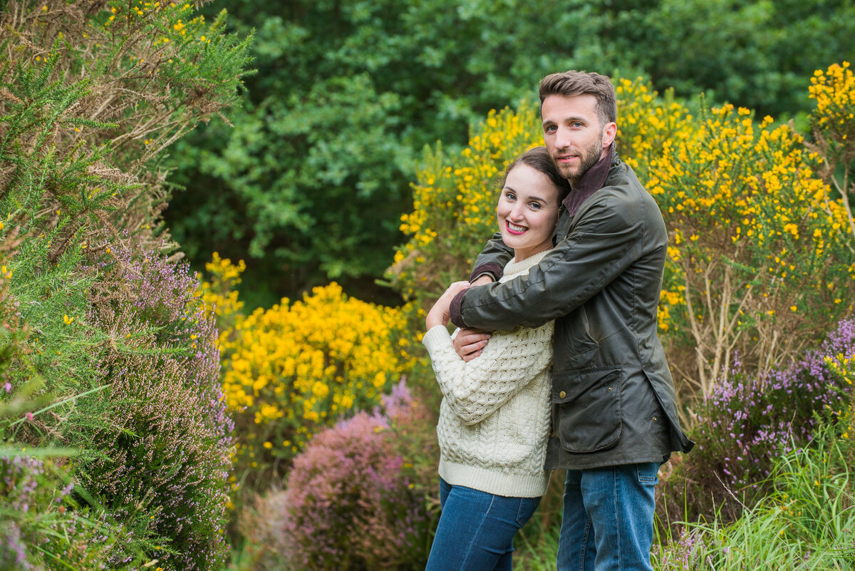 Young couple wearing an aran sweater and wax jacket with denim jeans hugging in a field, surrounded by flowers
