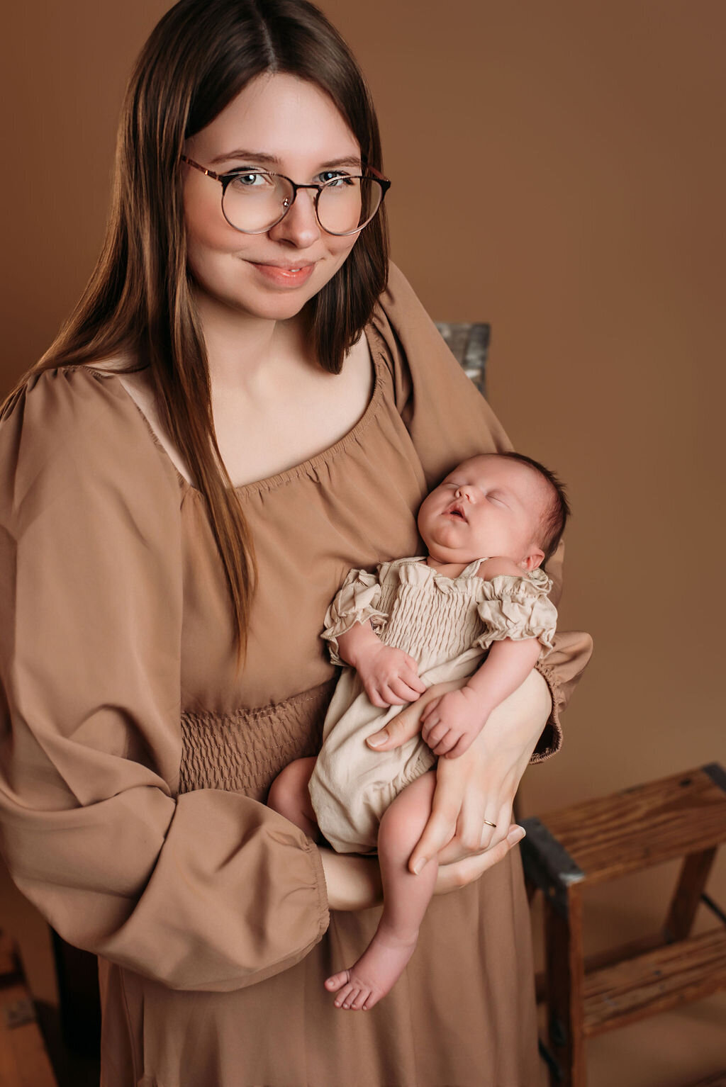 A woman in a tan boho dress sits in front of a neutral-toned backdrop holding a newborn baby in a beige outfit, captured by a photographer in Bemidji, Minnesota.